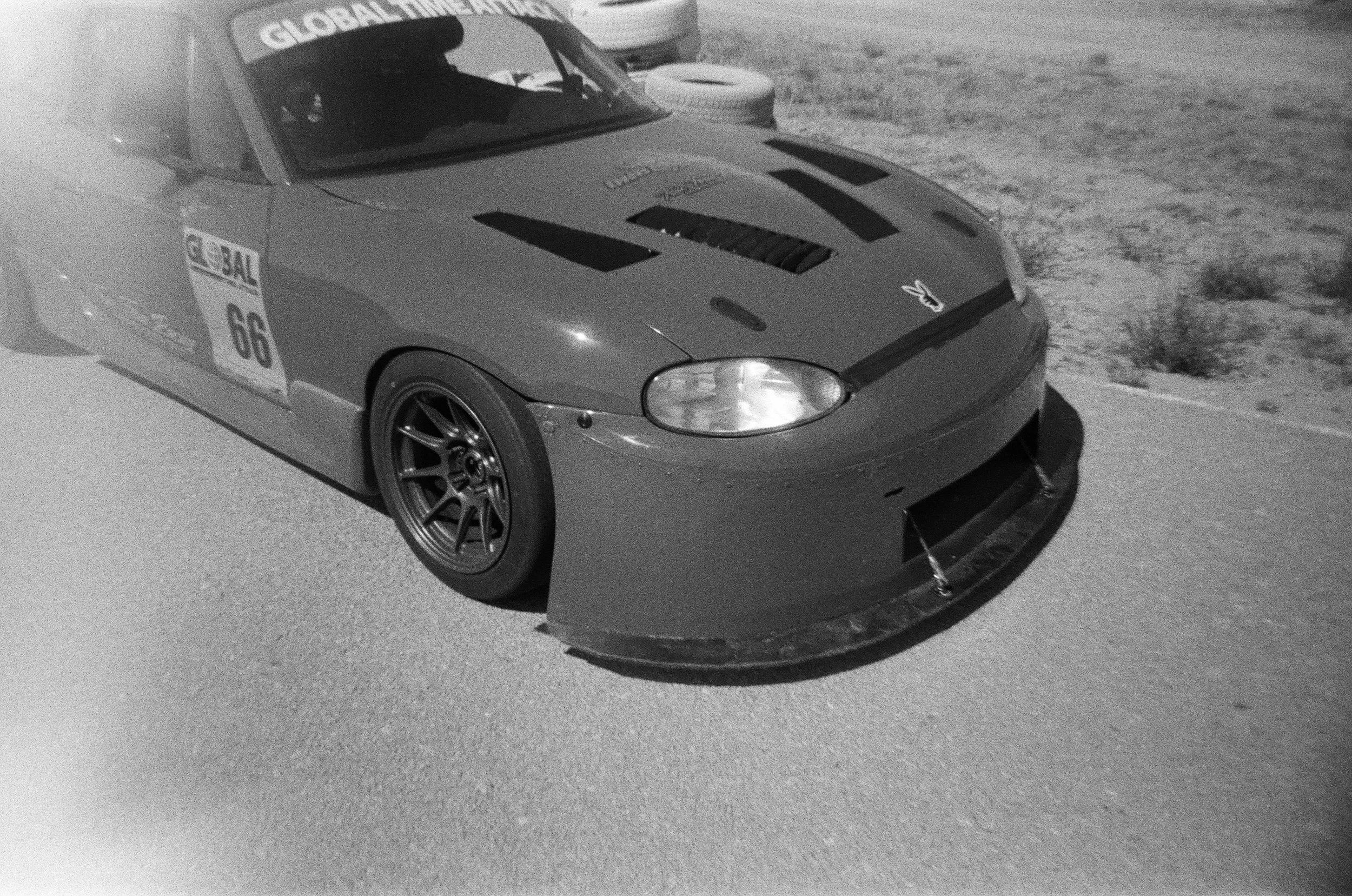 A race car, black with the number 66 on the side, parked on a desert road with a spare tire on the roof. The front splitter is extended and the car has various decals, including one that says 'Global Timing'.