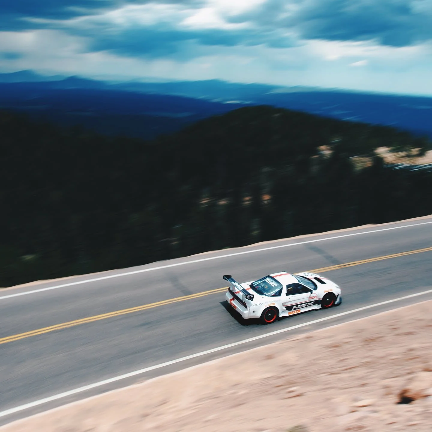 A white race car with black and red accents speeding down a mountain road with yellow dividing lines, blurred background of trees and mountains, and cloudy sky.
