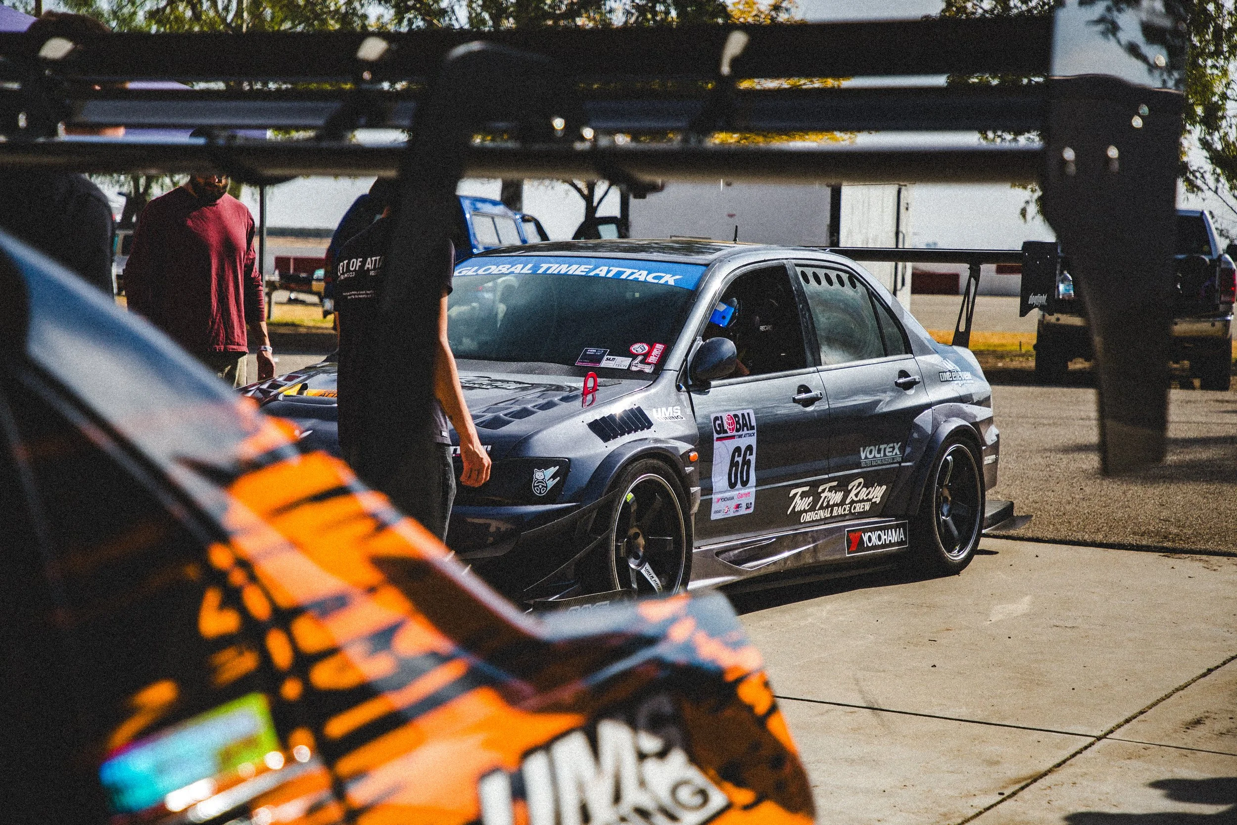 Race car parked under a shelter with people inspecting it, race team branding visible on the car, including 'Global Time Attack' and 'Yokohama' logos.