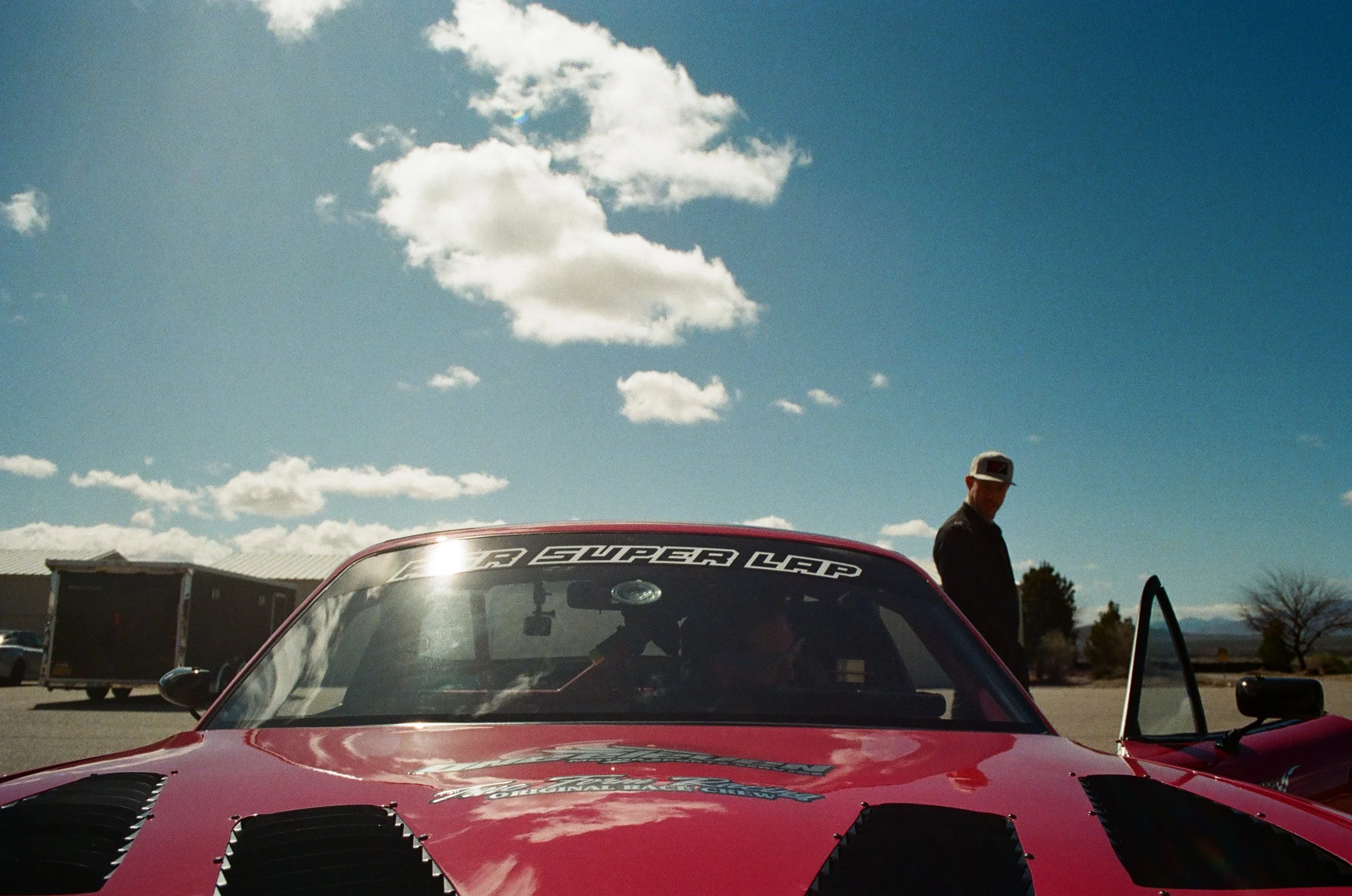 Red race car with a windshield banner that reads 'Super Lap' in front of a person standing outside under a blue sky with white clouds and a building in the background.