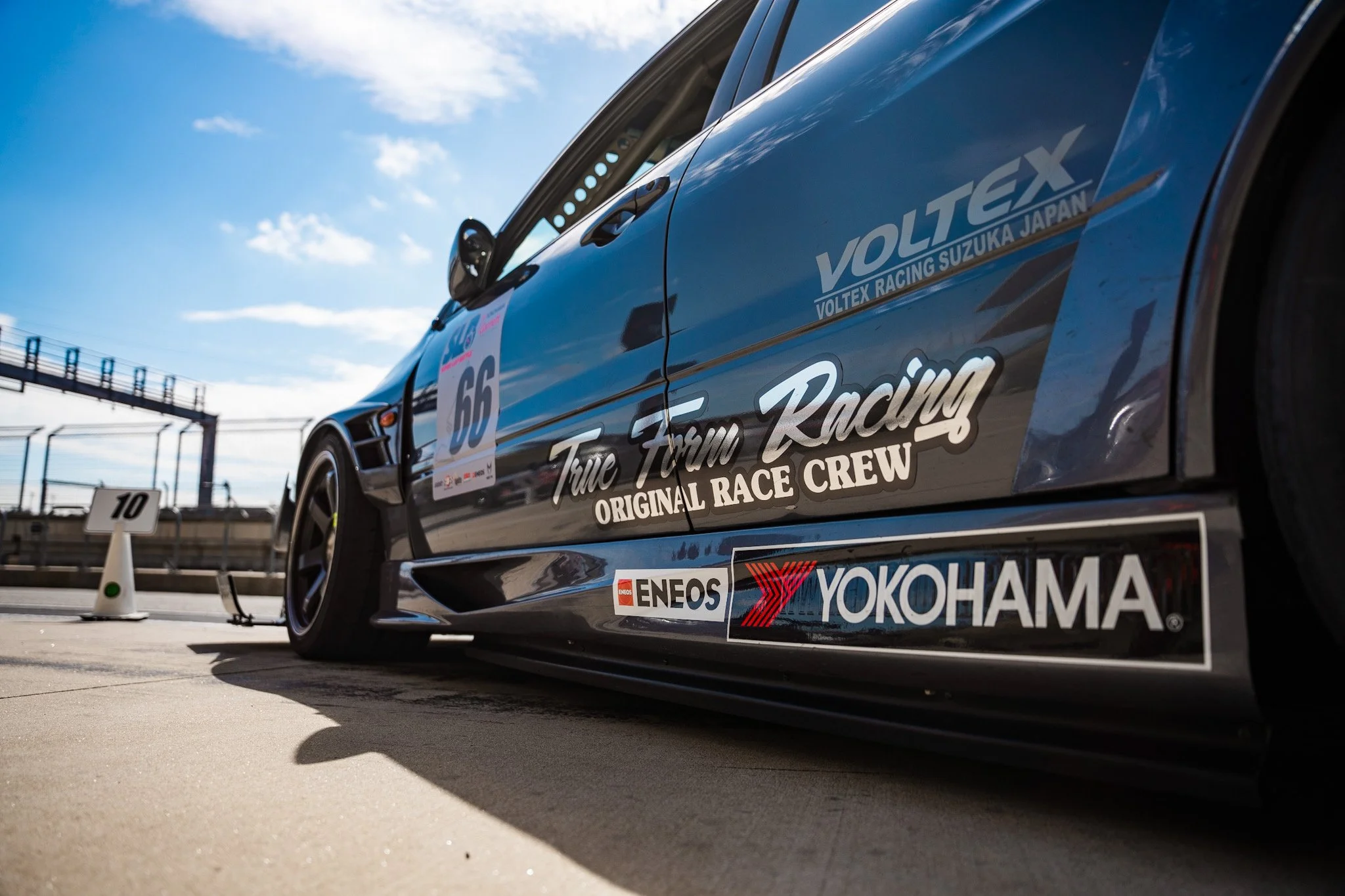 A race car on a track with sponsor logos, including Yokohama and ENEOS, under a partly cloudy sky. The car has racing decals and a number 66.
