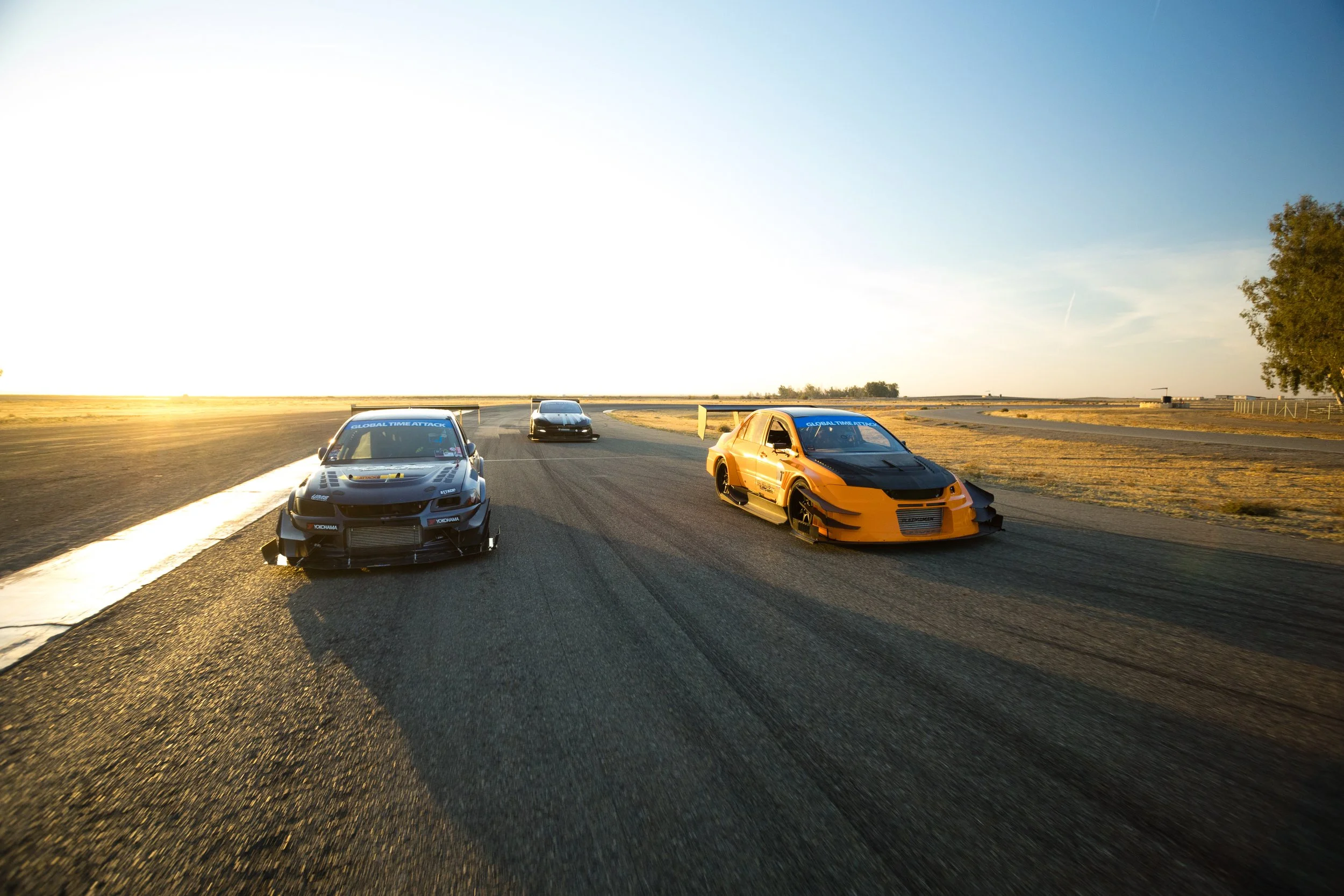 Four race cars driving on a track at sunset, with a landscape of open fields and trees in the background.