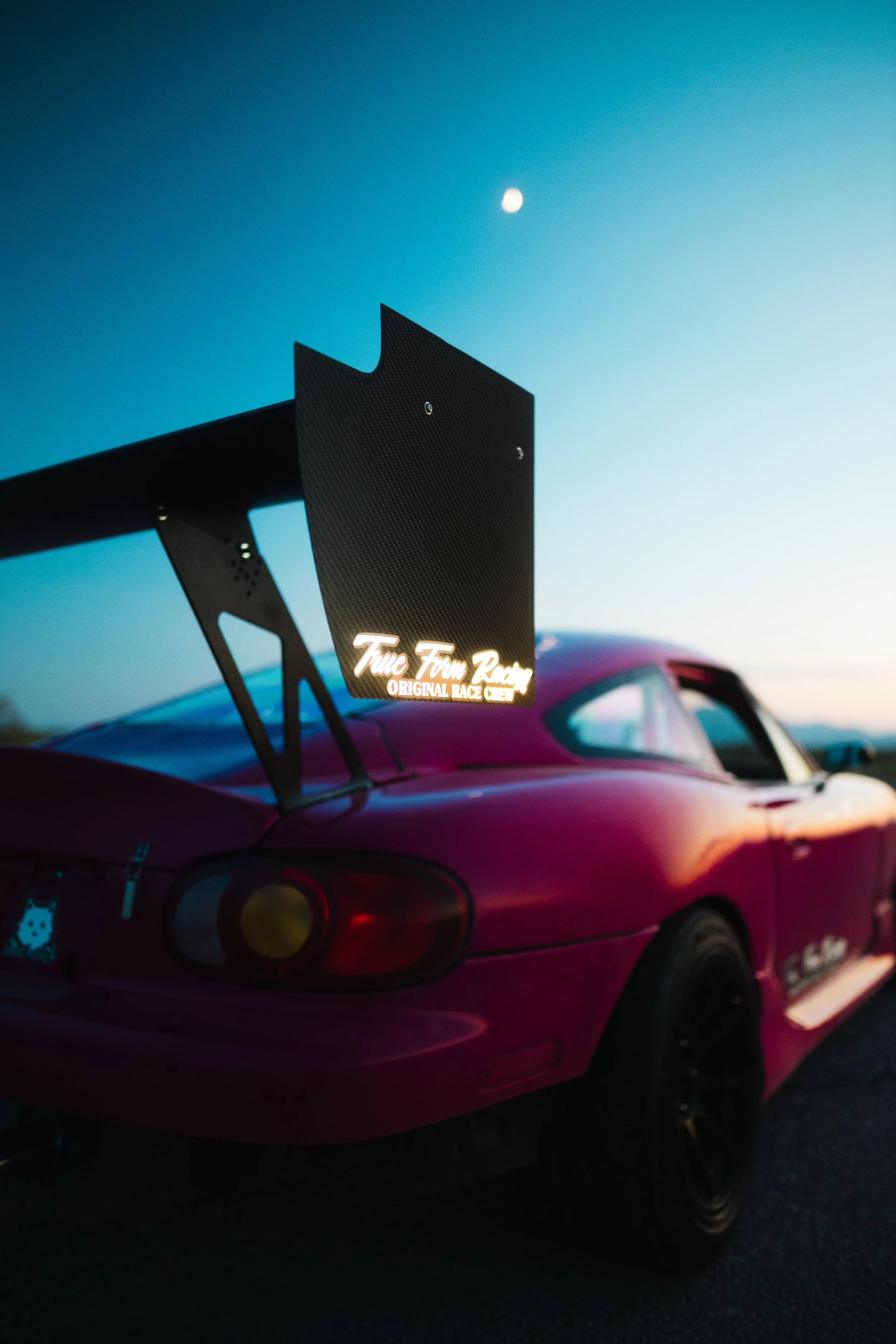 A pink sports car with stickers on the rear bumper and a large rear wing with a sign that reads 'True Tune Racing Original Race Team.' The background shows a clear sky with the moon visible. The car is parked on a dark surface.