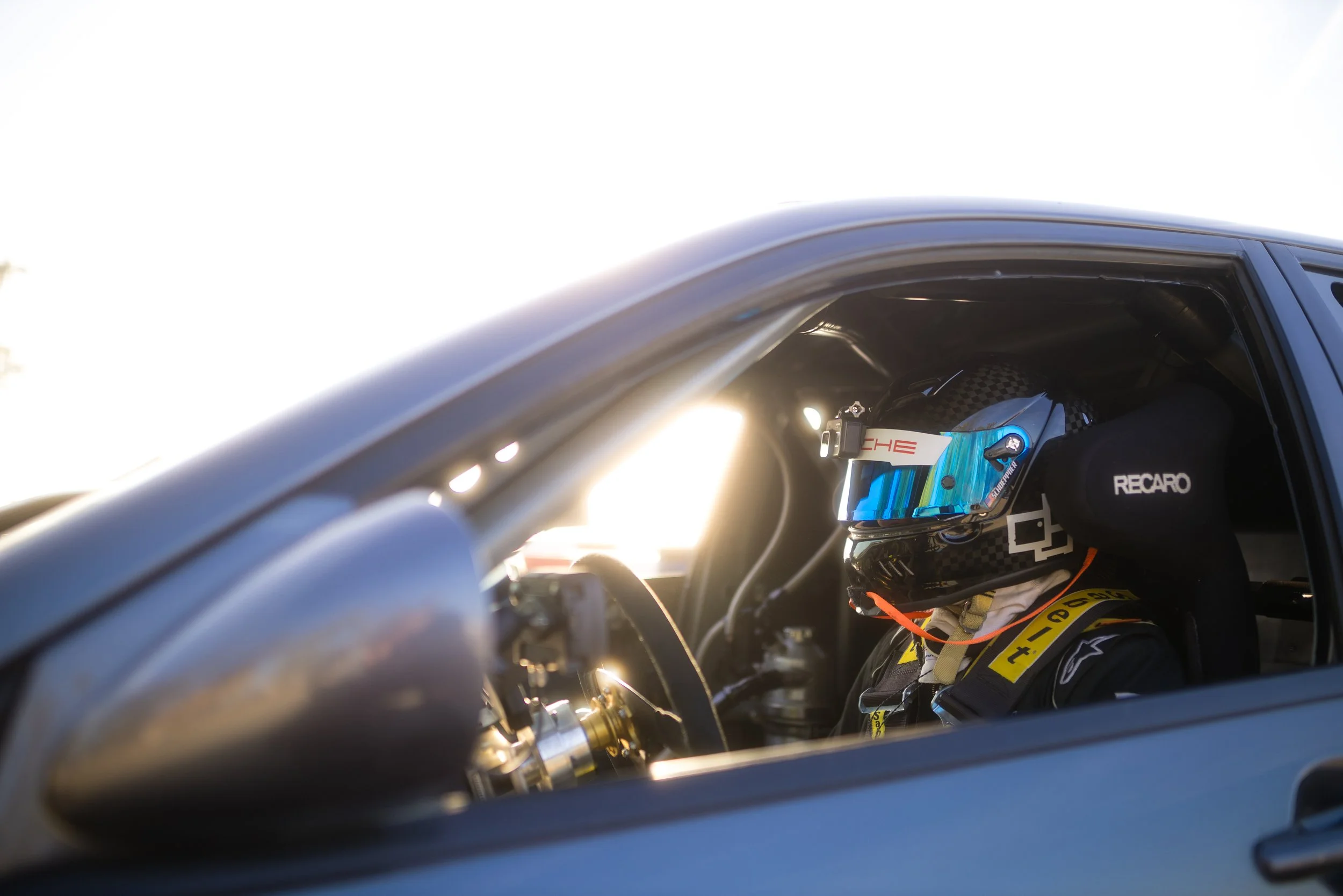 Race car driver wearing a black helmet with blue visor, seated inside a race car, with sunlight shining through the window and part of the car's interior visible.