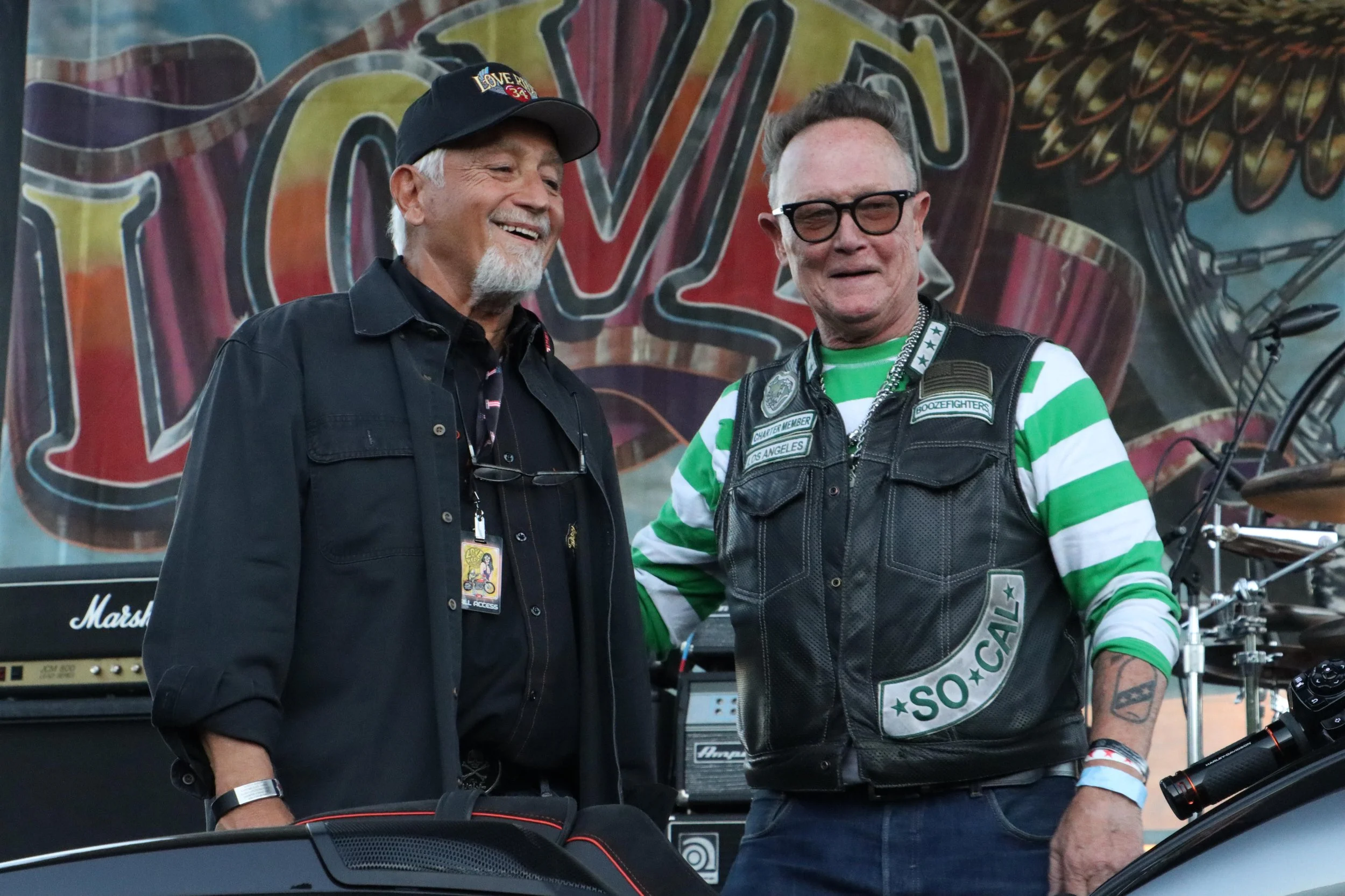 Two men smiling and talking at an event, with musical equipment and a large colorful backdrop behind them.
