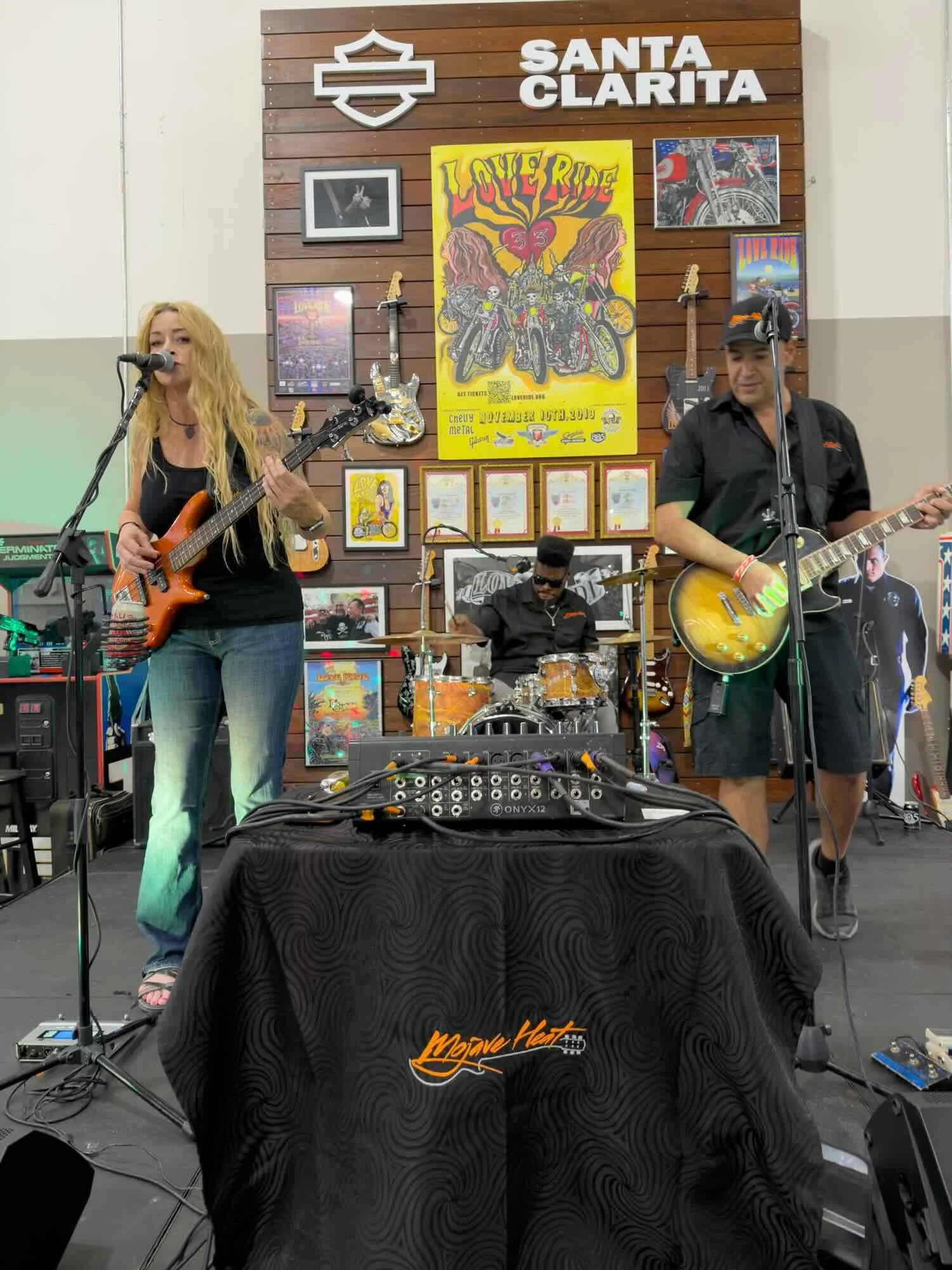 Band performing at a venue with a brick wall backdrop commemorating Santa Clarita, featuring guitars, posters, and a drummer. A woman with long blonde hair plays an electric guitar, and a man with a cap plays an acoustic guitar, while another man on drums is visible.
