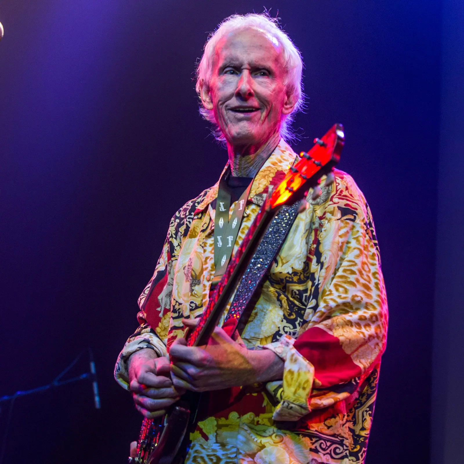 An elderly man with white hair playing a black electric guitar on stage, wearing a brightly colored, patterned shirt under purple and blue stage lighting.