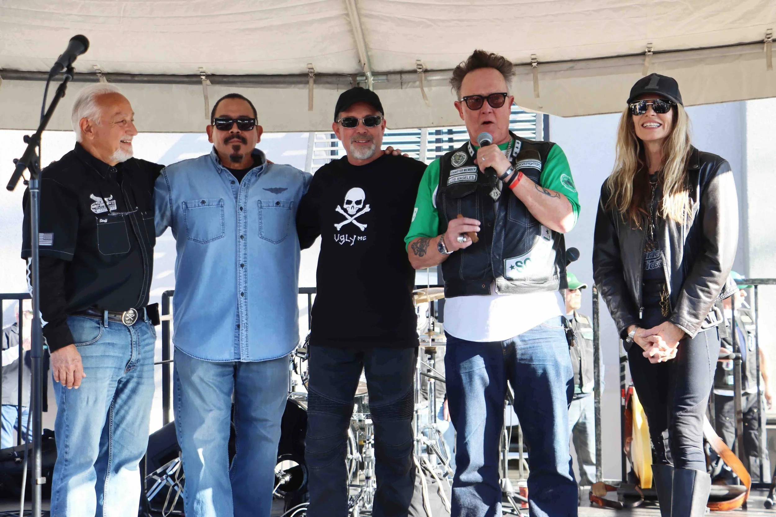 A group of five people standing under a tent, with some of them wearing sunglasses and casual attire, and one person speaking into a microphone.