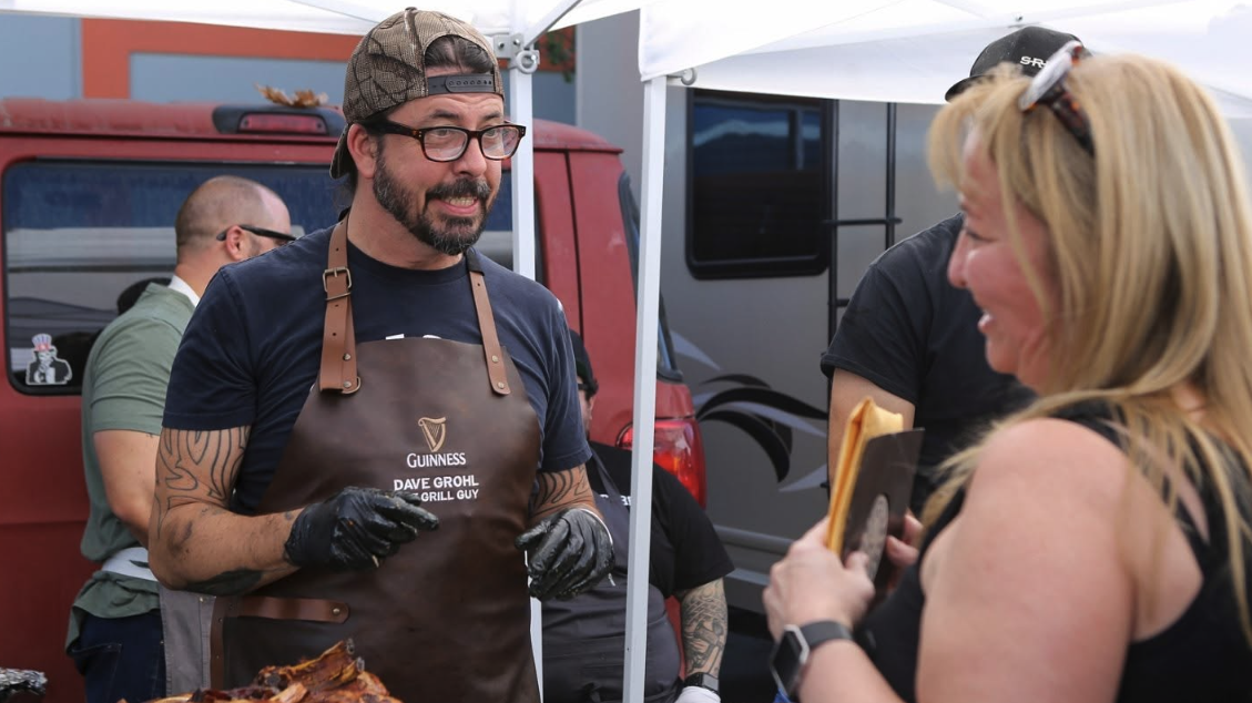 A man with glasses, a beard, and tattoos wearing a baseball cap, apron, and gloves talking to a woman holding a and McDonald's sandwich at an outdoor food event.