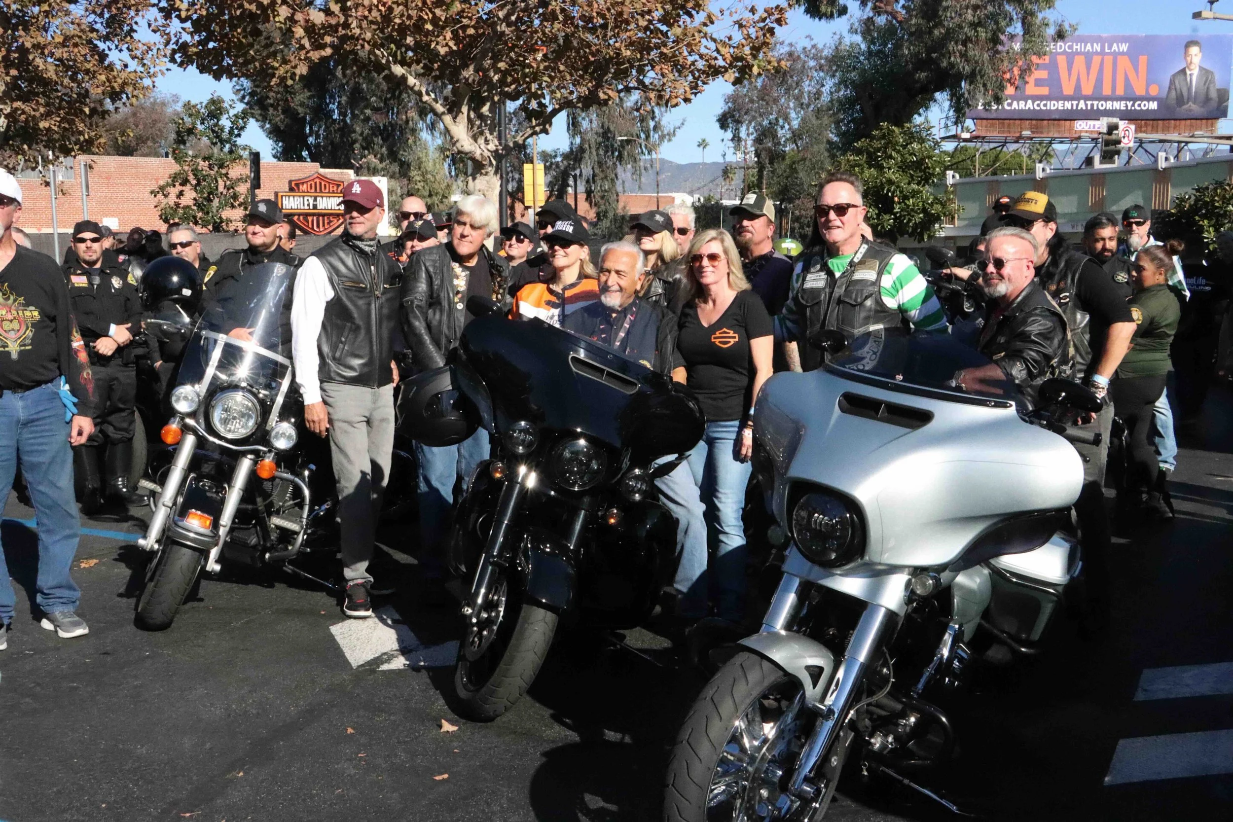 Group of people, including motorcyclists and civilians, gathered outdoors on a sunny day, with some wearing leather jackets and sunglasses, in front of motorcycles and a line of police officers. There are trees, a Harley-Davidson sign, and a billboard in the background.