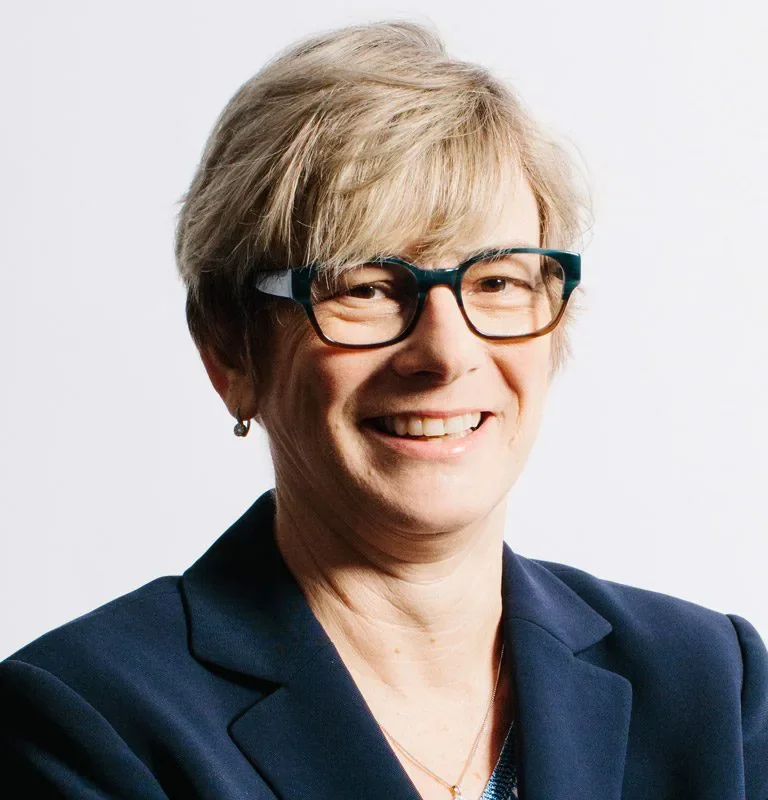 A middle-aged woman with short blonde hair, wearing glasses, a navy blazer, and a small earring, smiling against a plain white background.