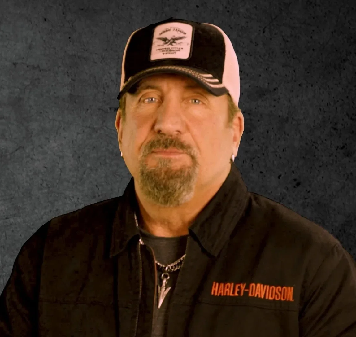 A man wearing a black and pink Harley-Davidson cap, black jacket with orange Harley-Davidson logo, with light-colored eyes, facial hair, and a chain necklace, standing against a dark textured background.