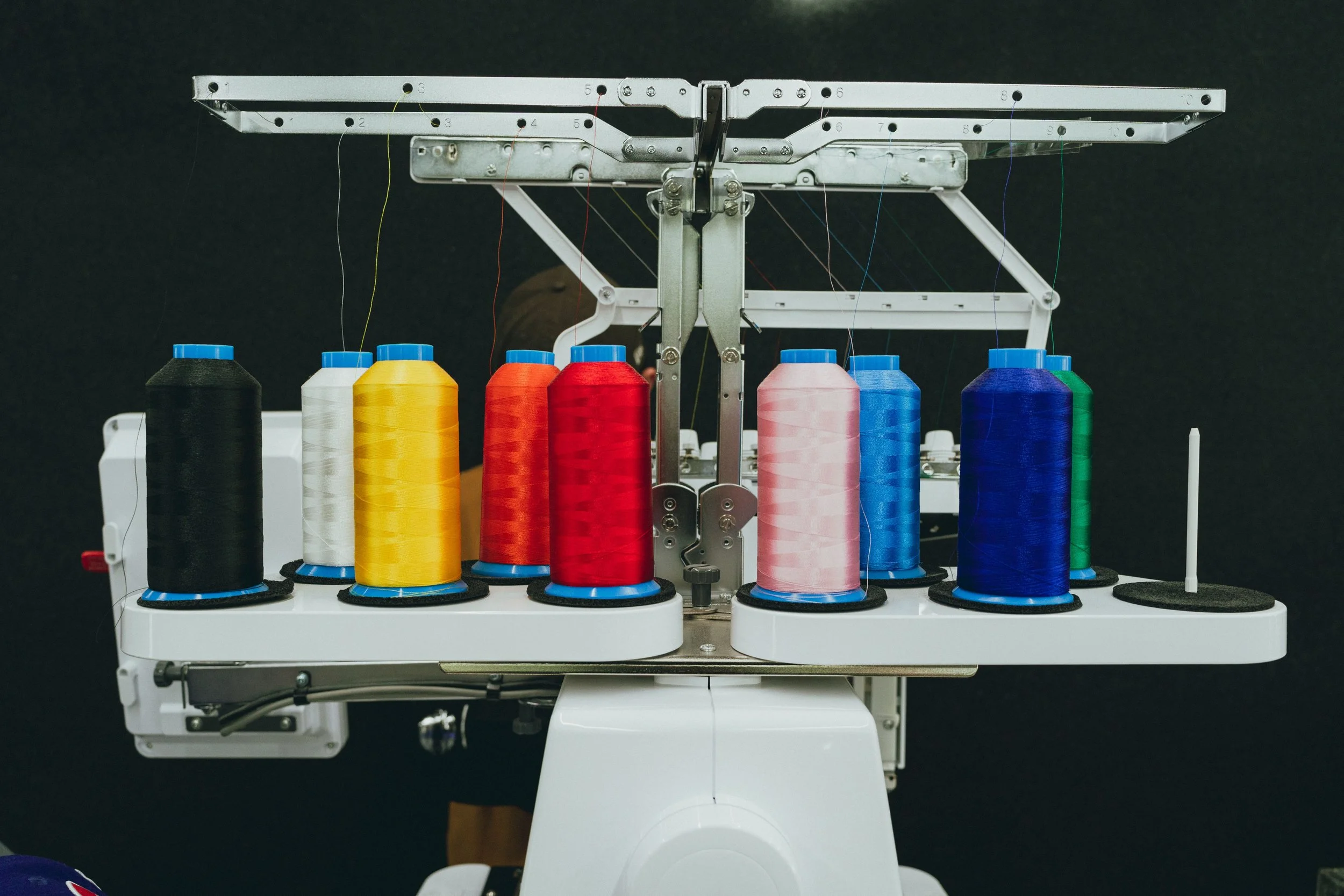 A computerized embroidery sewing machine with multiple spools of thread in black, white, yellow, red, pink, blue, and green. The machine is set against a black background.