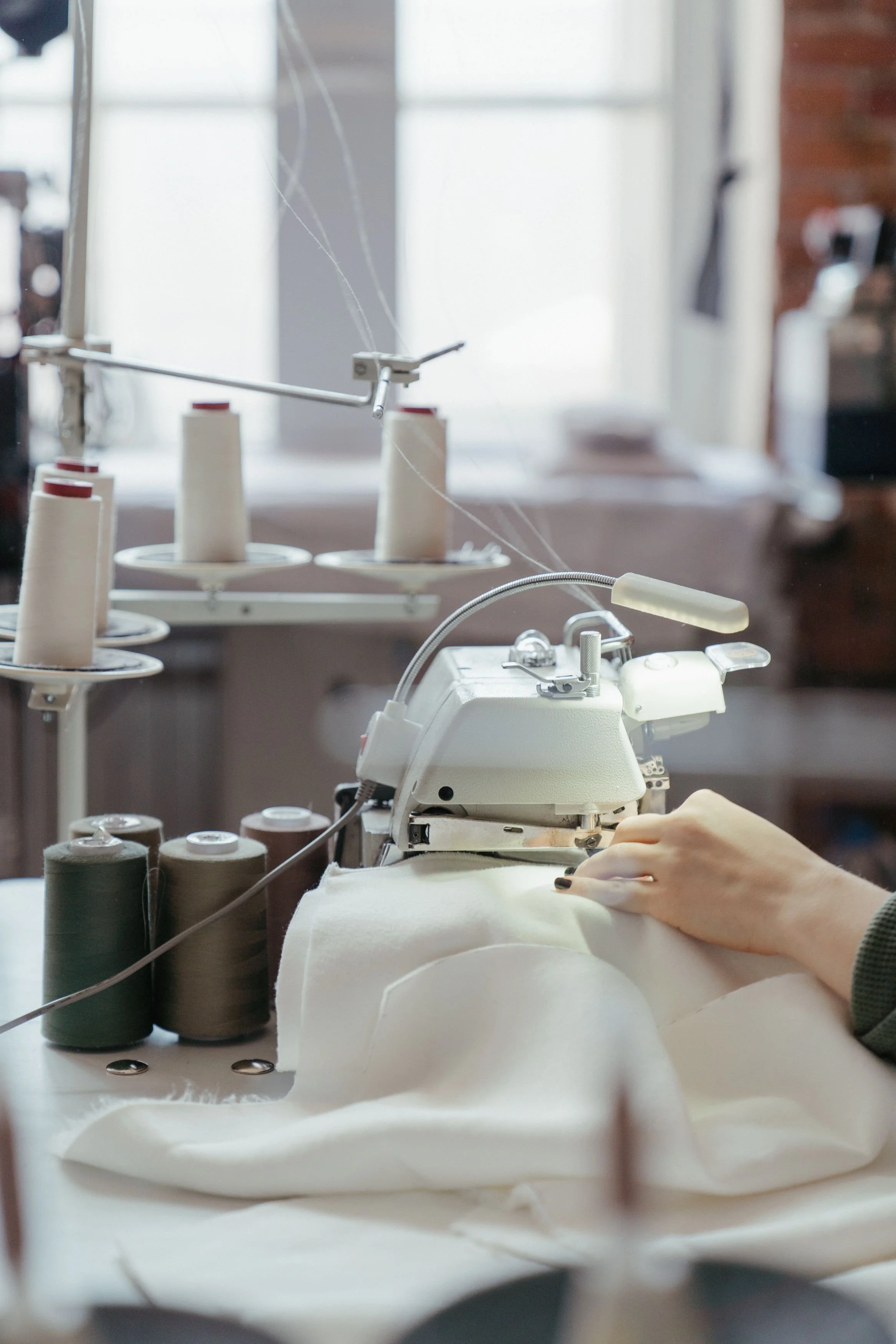 Close-up of a sewing machine with stacked spools of thread in different colors and a person's hand operating the machine, set on a worktable in a workspace.
