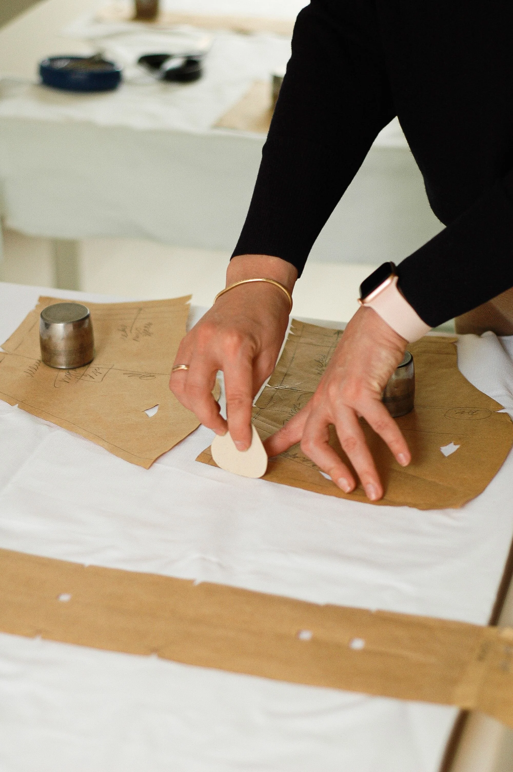 Person wearing a black long-sleeve top with a pink watch and gold bracelet, handling paper and metal objects on a white table, with a background of miscellaneous items.