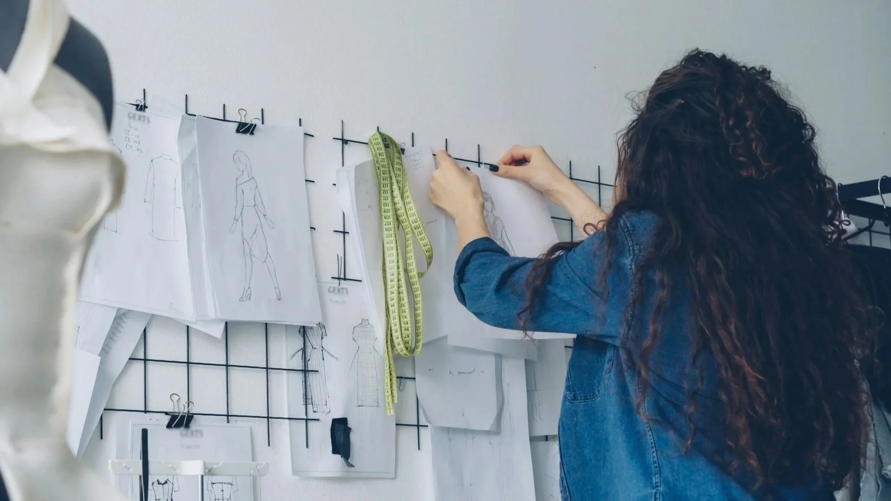 A woman with curly hair wearing a denim jacket pins or adjusts fashion sketches on a grid wall, with a yellow measuring tape hanging next to the sketches.