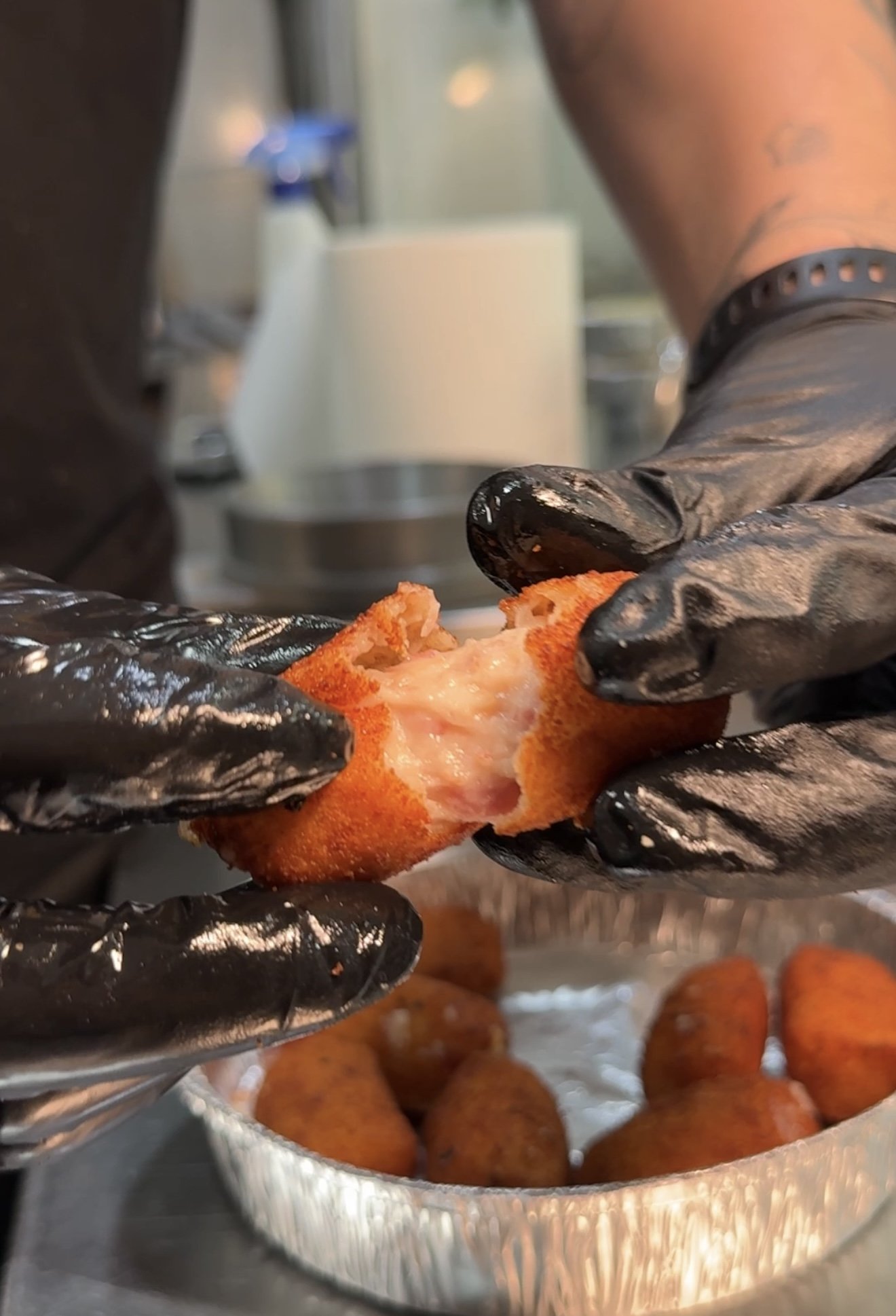Persona usando guantes negros preparando croquetas en un recipiente de aluminio.