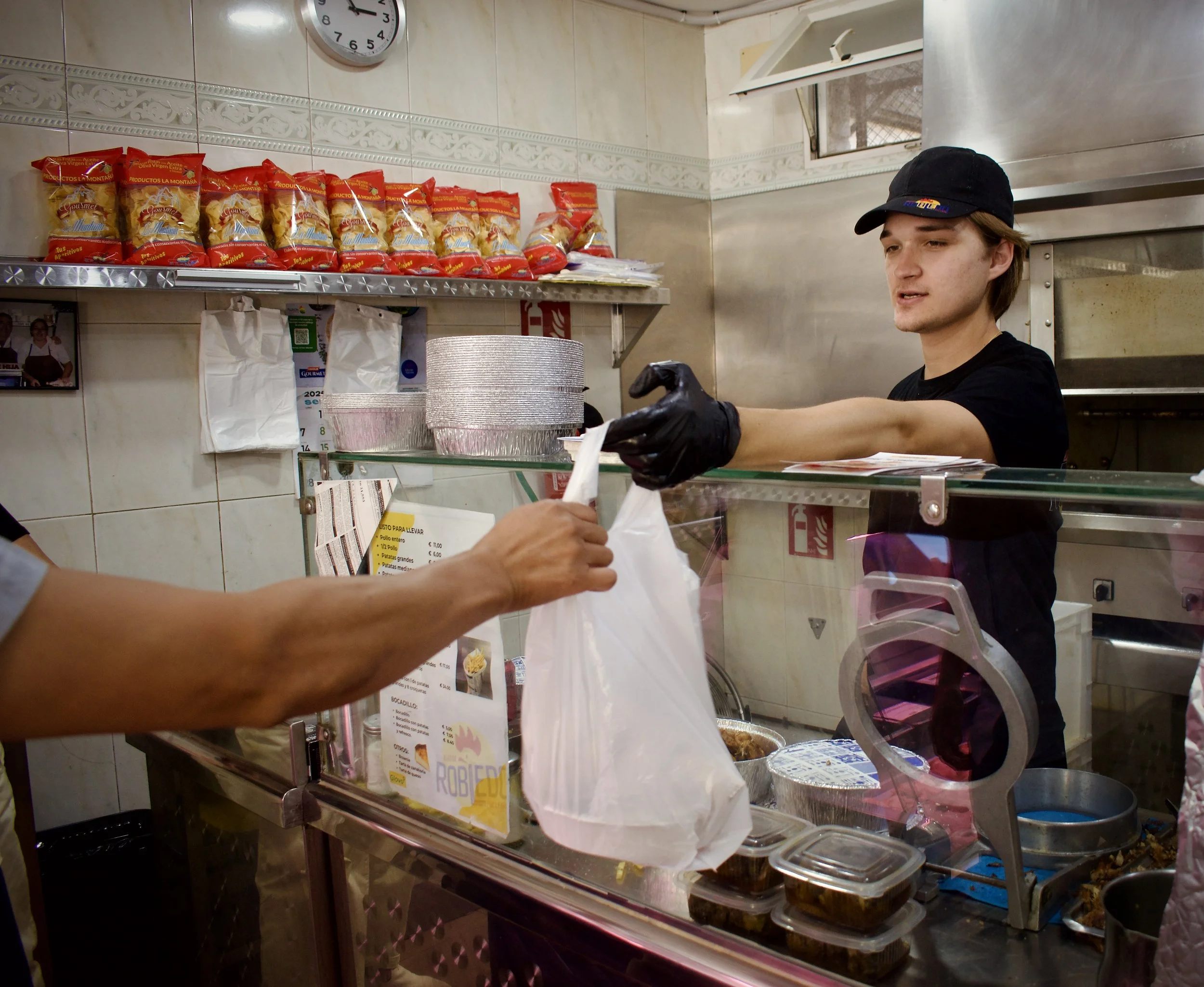 Joven atendiendo en una tienda de comida rápida, entregando un pedido en una bandeja con comida, detrás hay estantes con paquetes de pasta y un reloj en la pared.