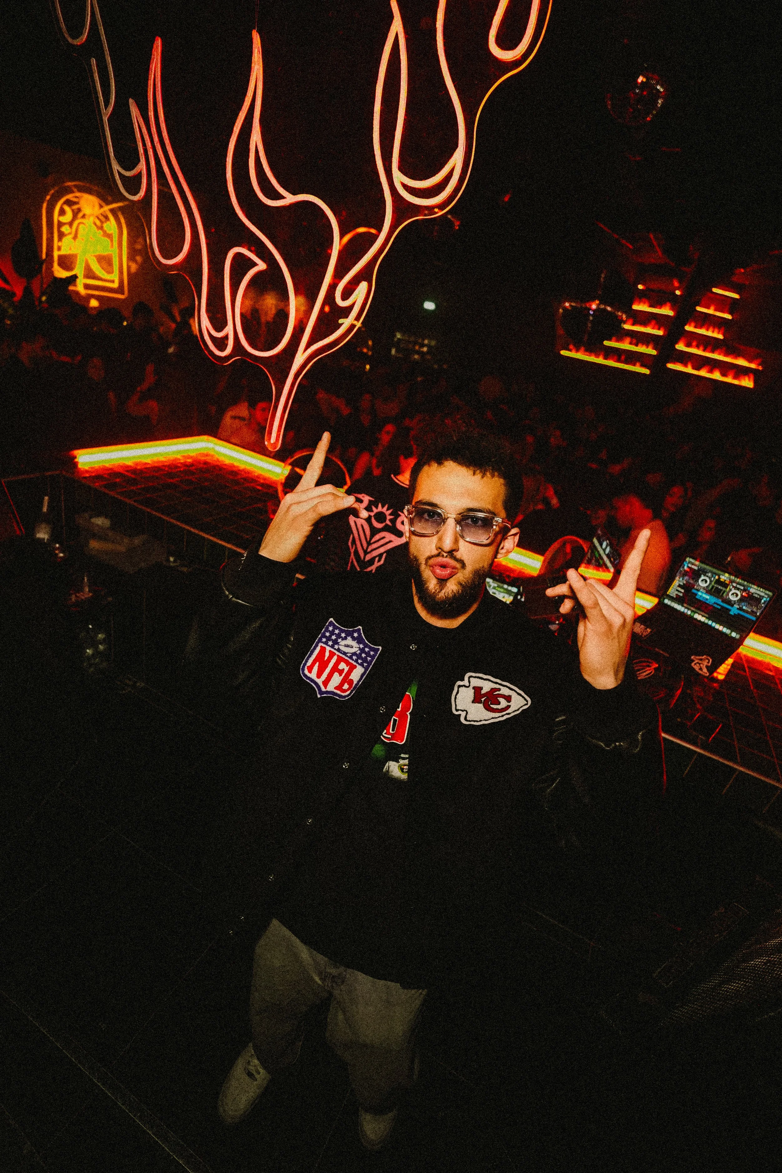 A person with glasses and a beard making a peace sign with both hands, in a dimly lit nightclub or bar with neon lights and a DJ setup in the background. The person is wearing a jacket with NFL patches.