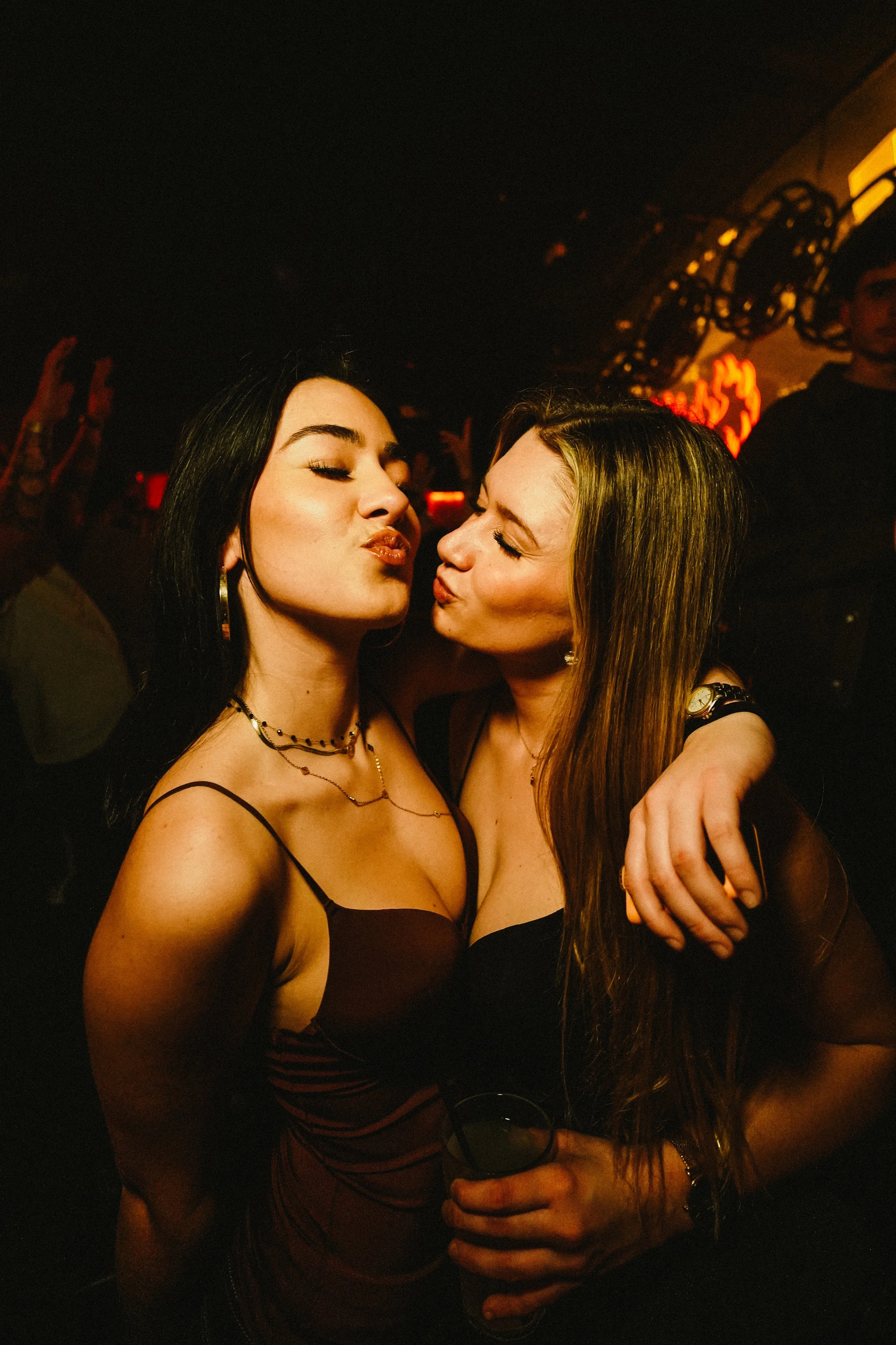 Two women are close together at a party, kissing each other and smiling. They are wearing dark dresses and jewelry, and one is holding a glass of wine.