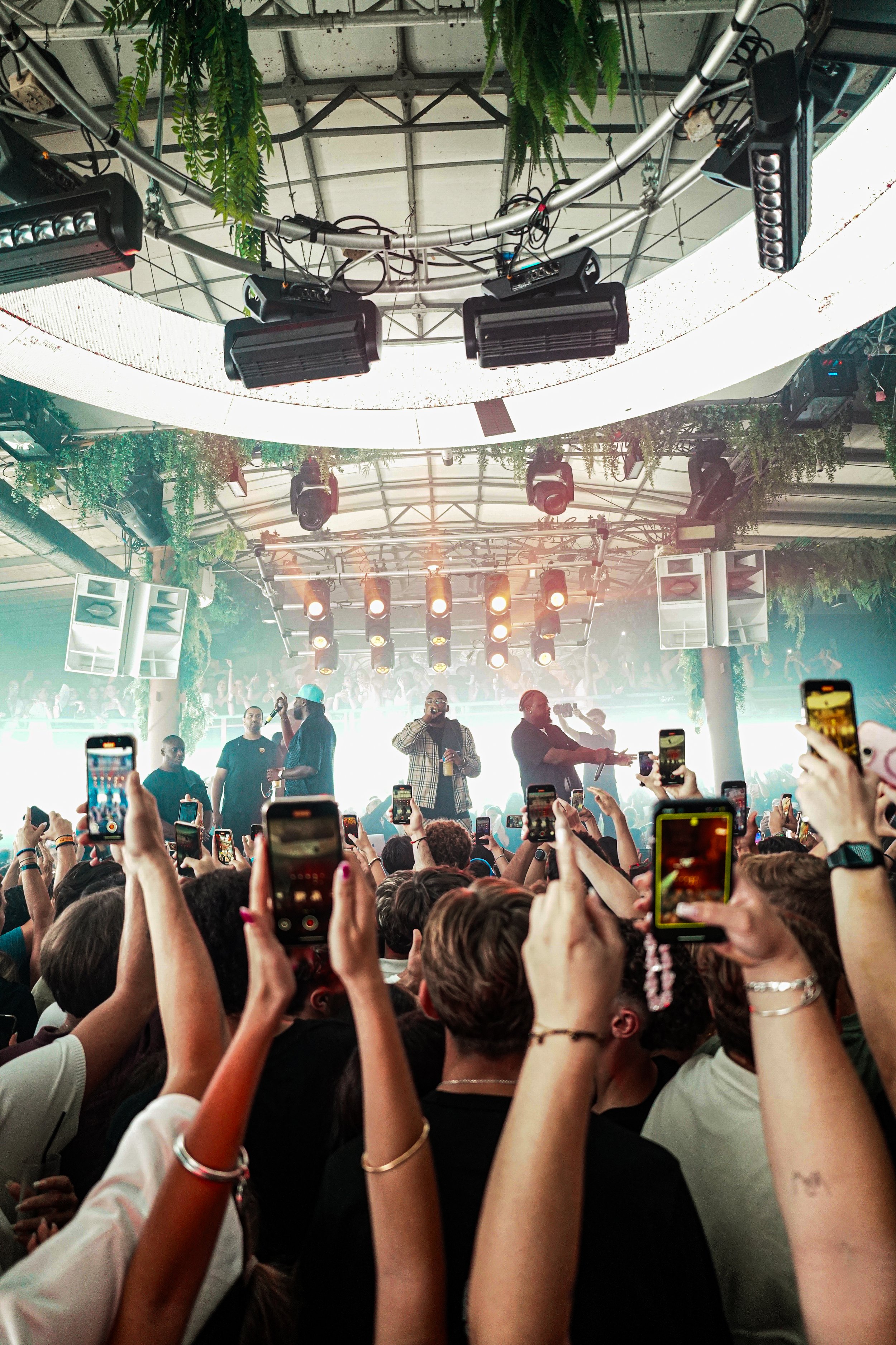 Crowd of people at a concert or event with hands raised, many holding smartphones, stage with performers under hanging lights and greenery, ceiling with lighting and audio equipment.