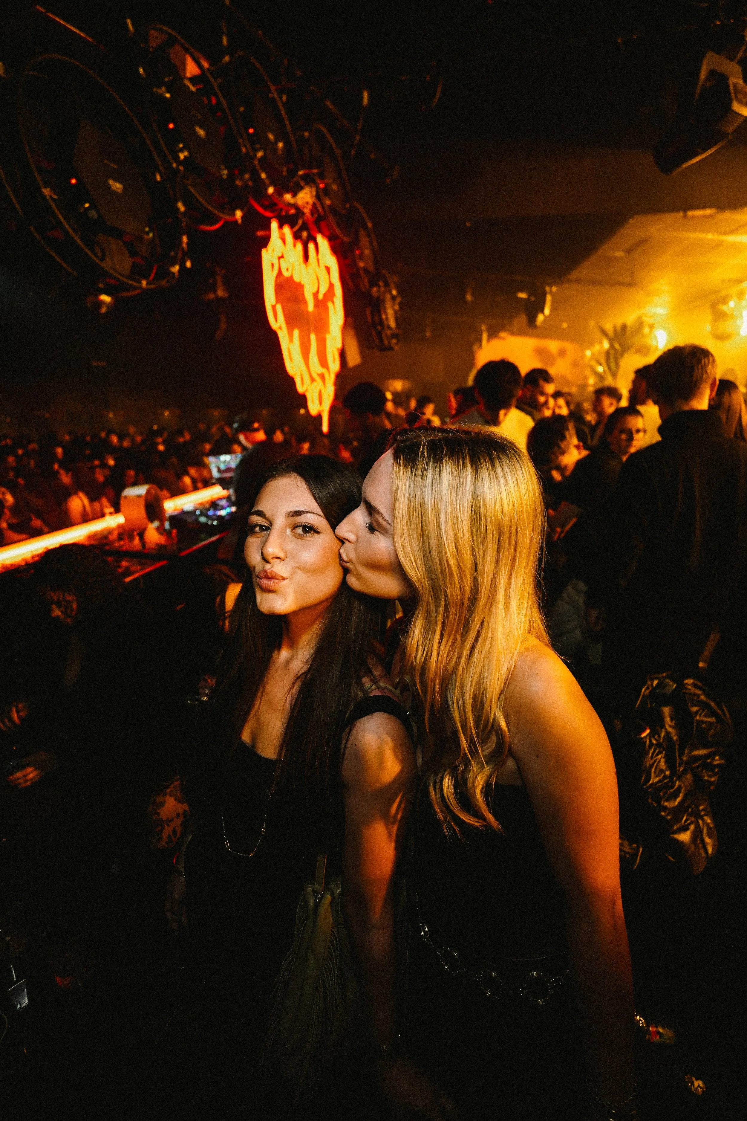 Two women are taking a selfie in a crowded nightclub with warm, orange lighting. One woman is puckering her lips, and the other is kissing her cheek. In the background, there are many people, DJ equipment, and a neon sign.