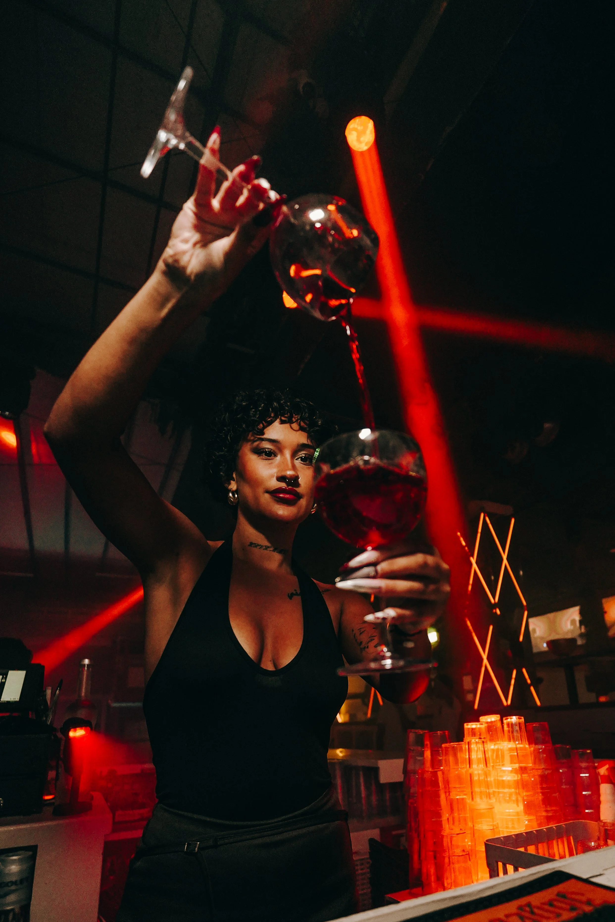A woman with curly hair, tattoos, dark lipstick, and earrings pouring red wine into a glass at a bar with orange lighting and stacked plastic cups.