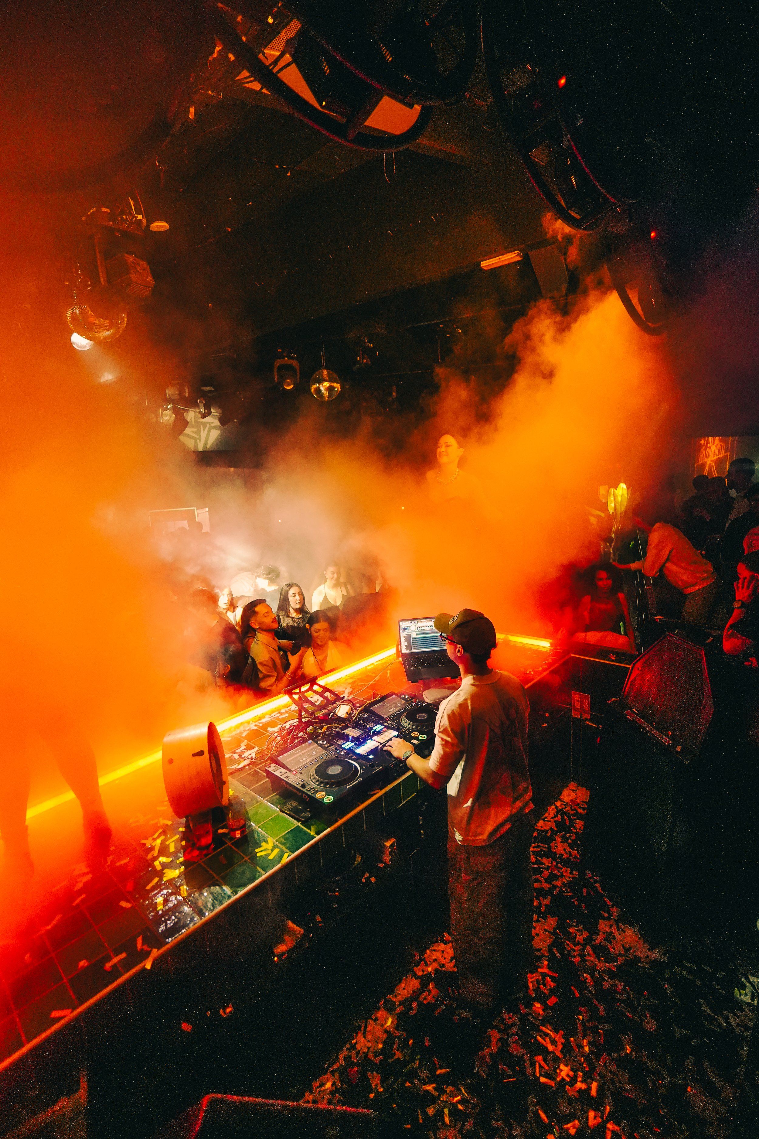 A DJ performing at a nightclub with a crowd dancing. The scene is filled with orange fog, colorful lighting, and disco balls hanging from the ceiling.