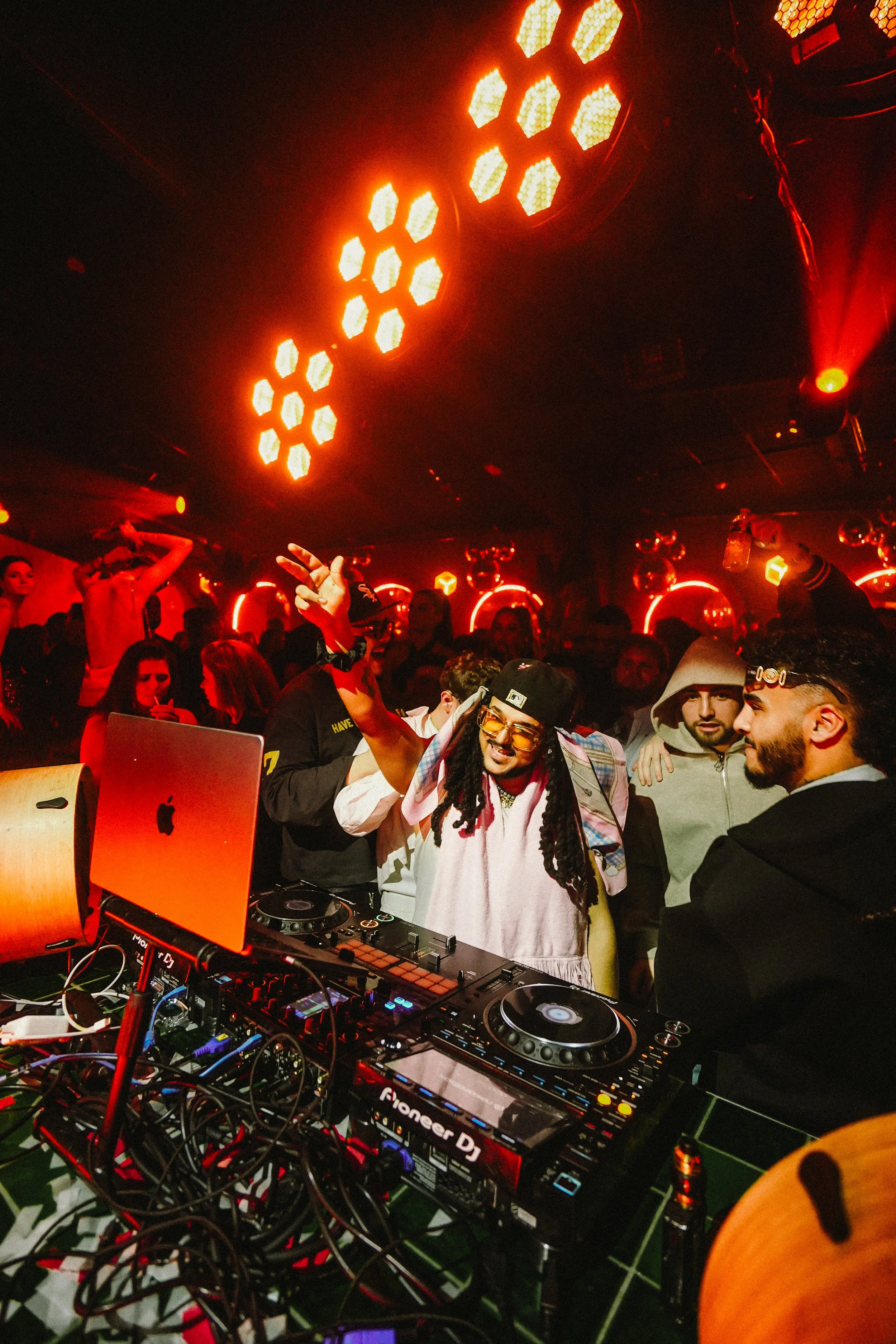 Nightclub scene with a DJ wearing glasses and a hat, playing music behind DJ equipment, surrounded by people dancing and socializing. Red lighting and disco balls decorate the ceiling.
