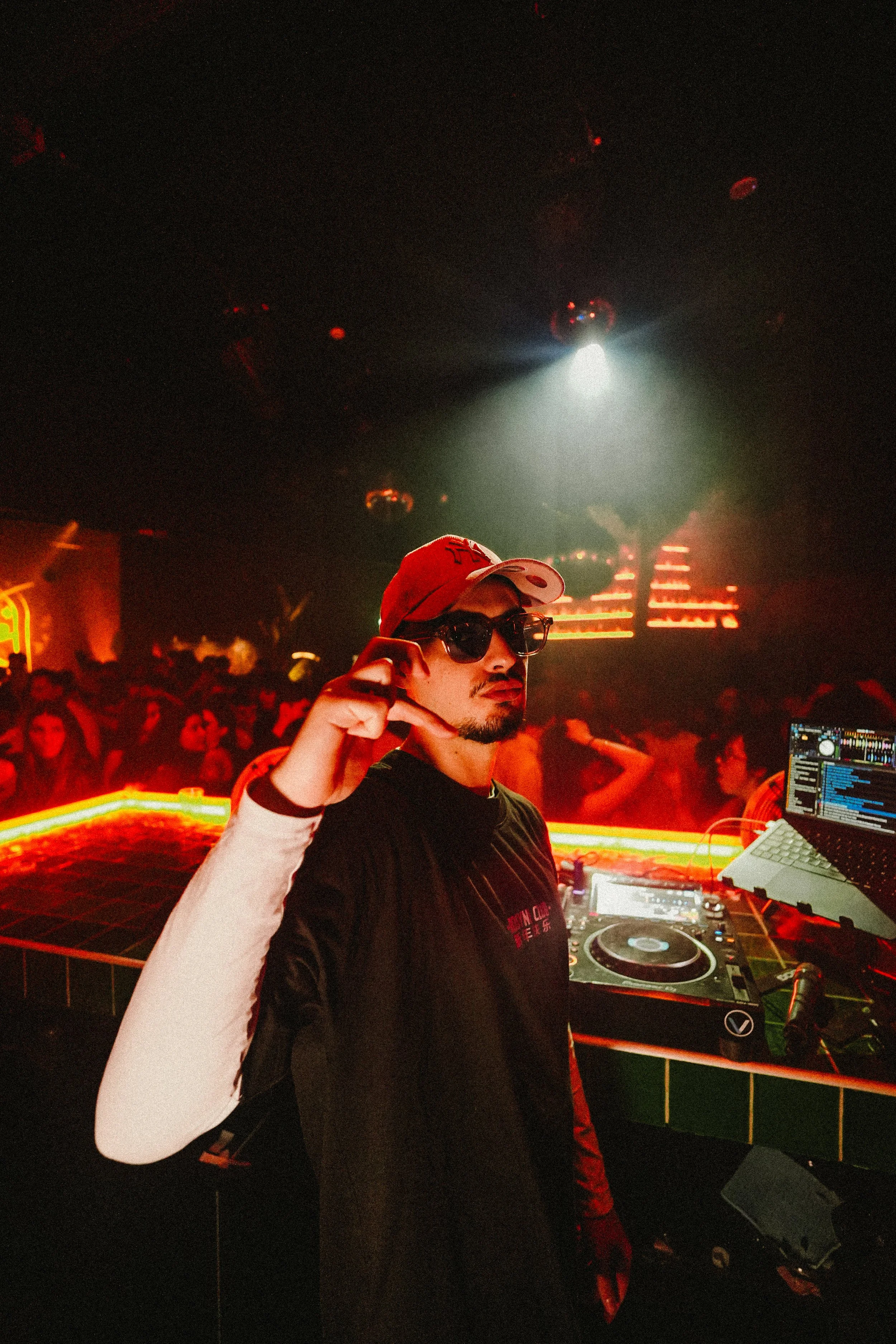 A DJ wearing sunglasses and a red cap, standing behind a DJ booth with turntables and audio equipment in a nightclub with a crowd and colorful lighting.