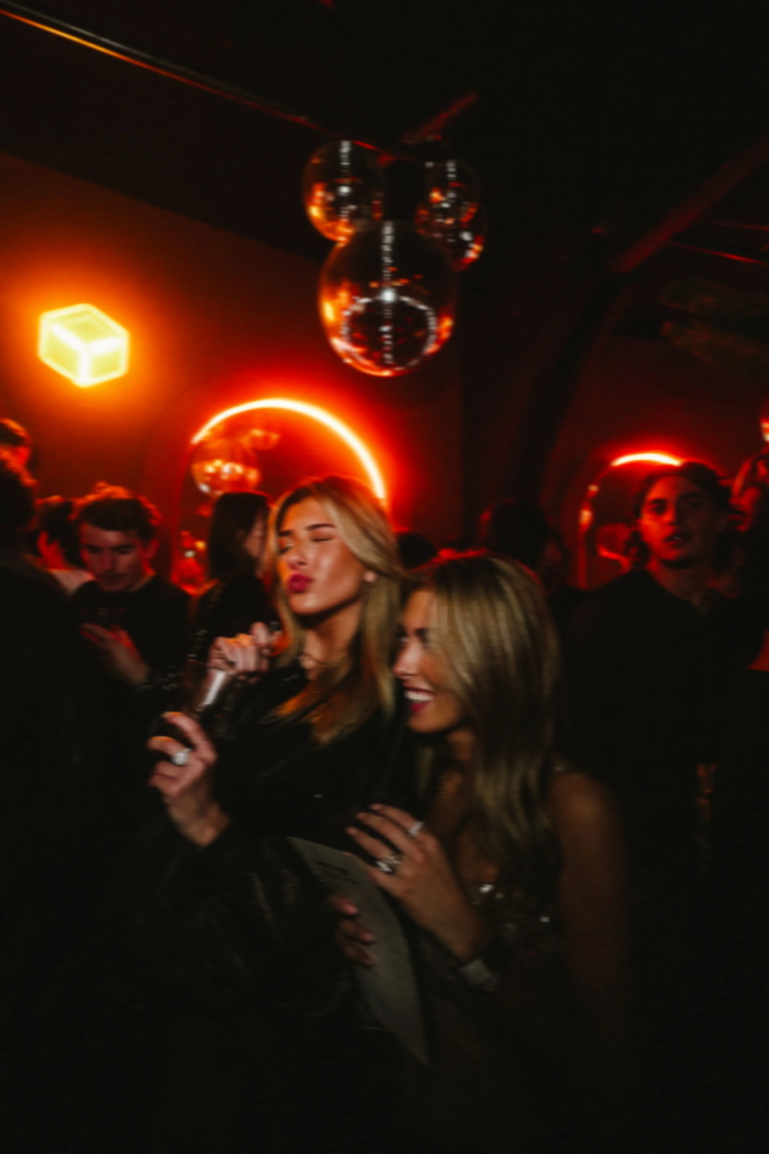 Group of people dancing and socializing in a dark nightclub with red and orange lighting and hanging disco balls.