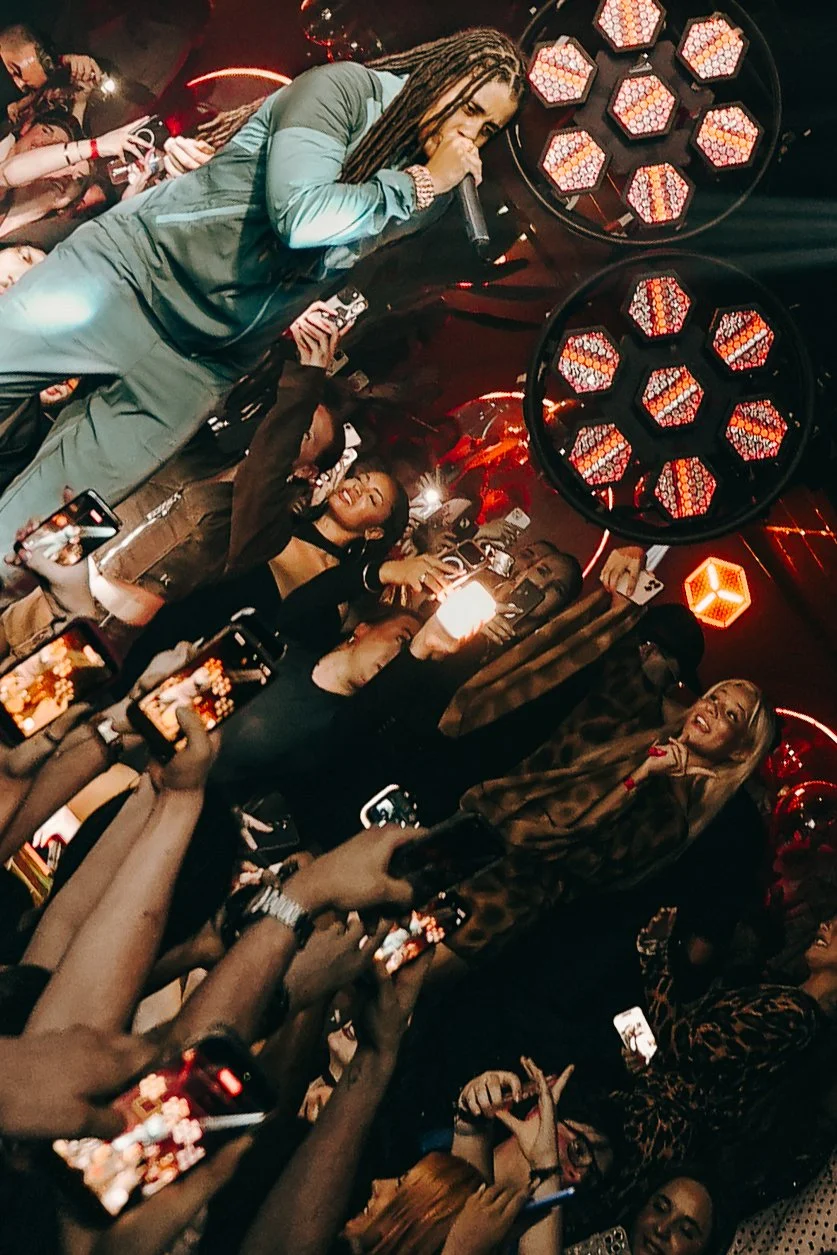 A performer with long dreadlocks, wearing a green jacket, sings into a microphone on stage during a lively event. The crowd around her is holding up phones to record or take pictures, with red and orange lighting creating an energetic atmosphere.