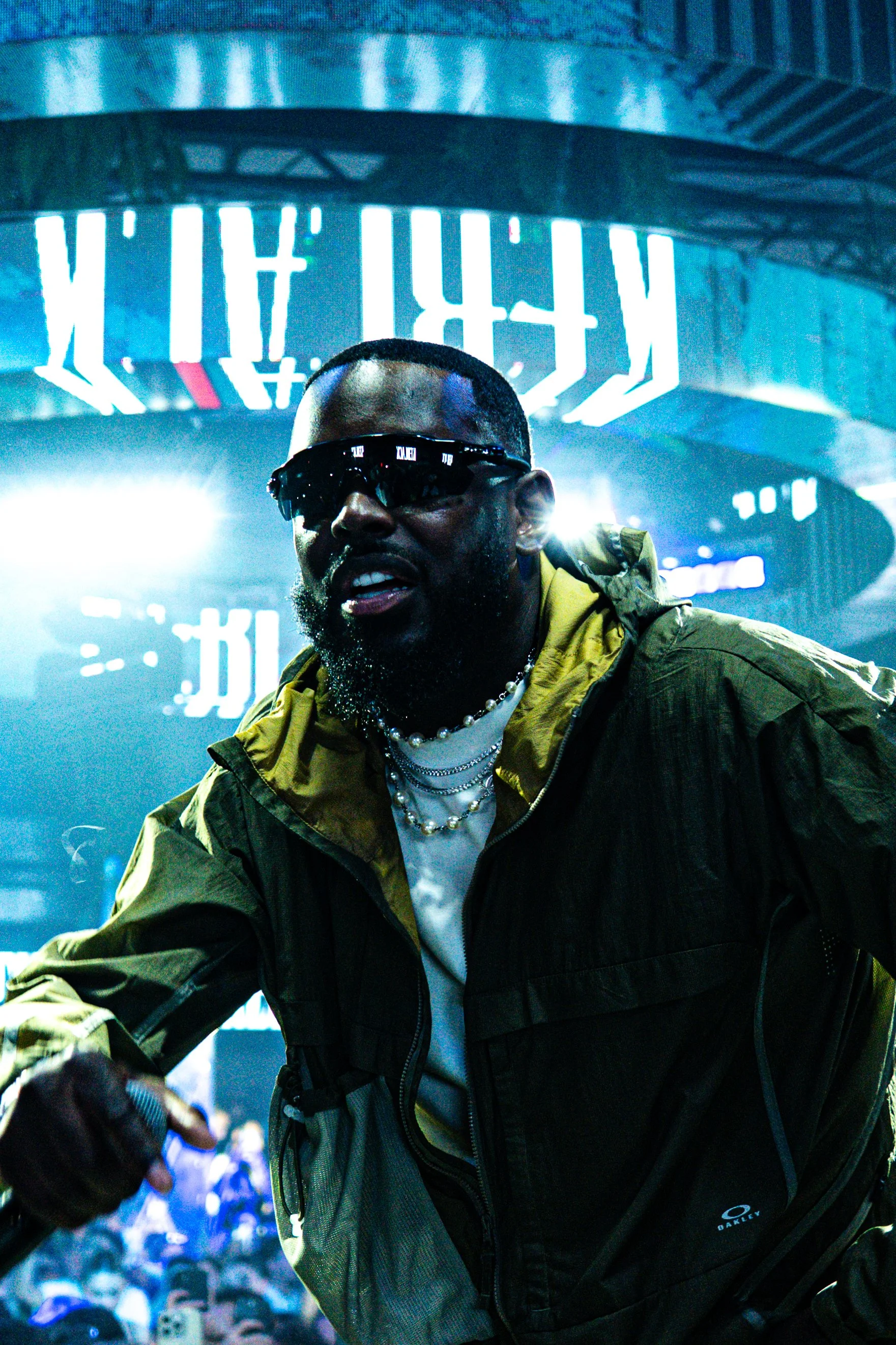 A man wearing sunglasses, pearl necklaces, and a yellow and black Oakley jacket captures a vibrant night scene with bright lights and Chinese characters in the background.