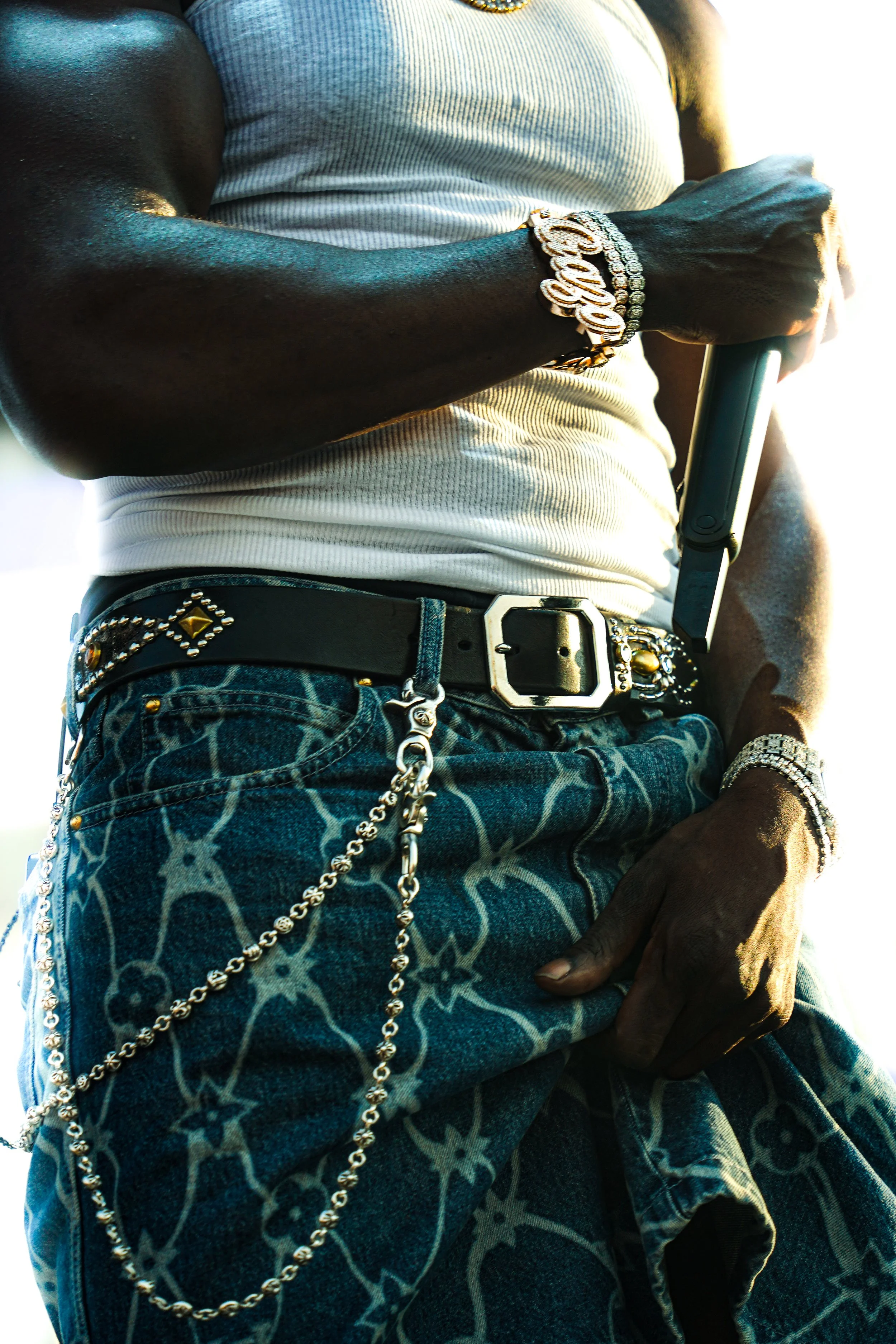 Close-up of a man's arm, wearing a gray tank top, multiple jewelry including a bracelet with the word 'Love,' a watch, and a chain. He is also wearing patterned jeans with chains and accessories. The background is bright with sunlight.