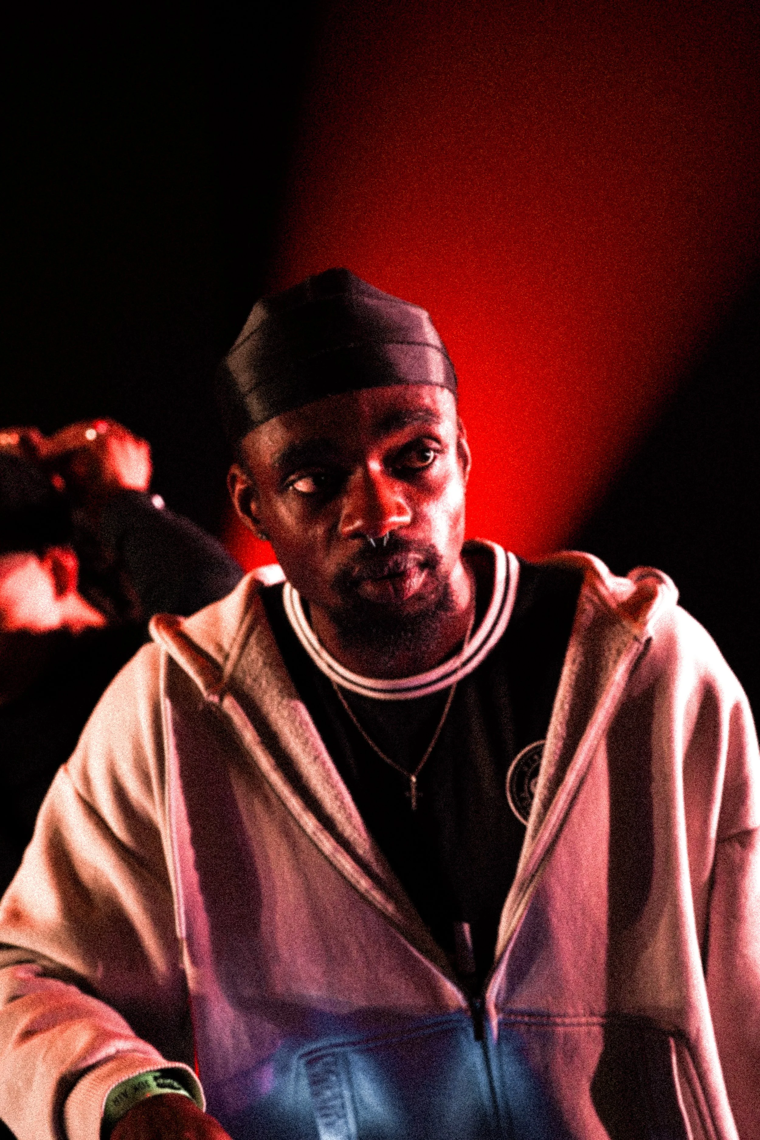 A man with dark skin wearing a black beanie cap, a black and white jacket, and a black shirt, smiling slightly in a dimly lit environment with red and black background lighting.