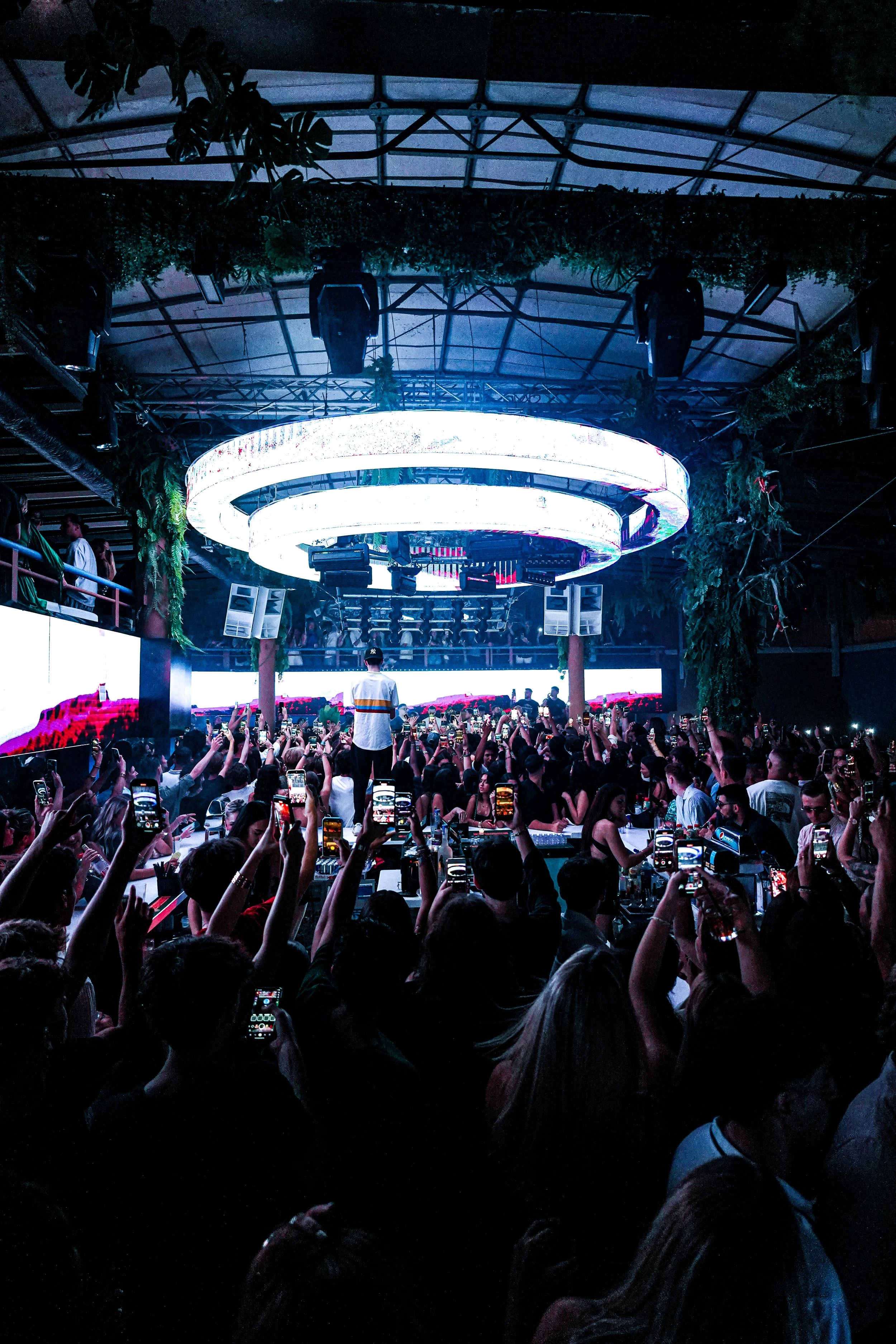 Crowd of people attending a live music event with a DJ performing under a large circular LED light fixture, in an indoor venue with a high ceiling and greenery decorations.