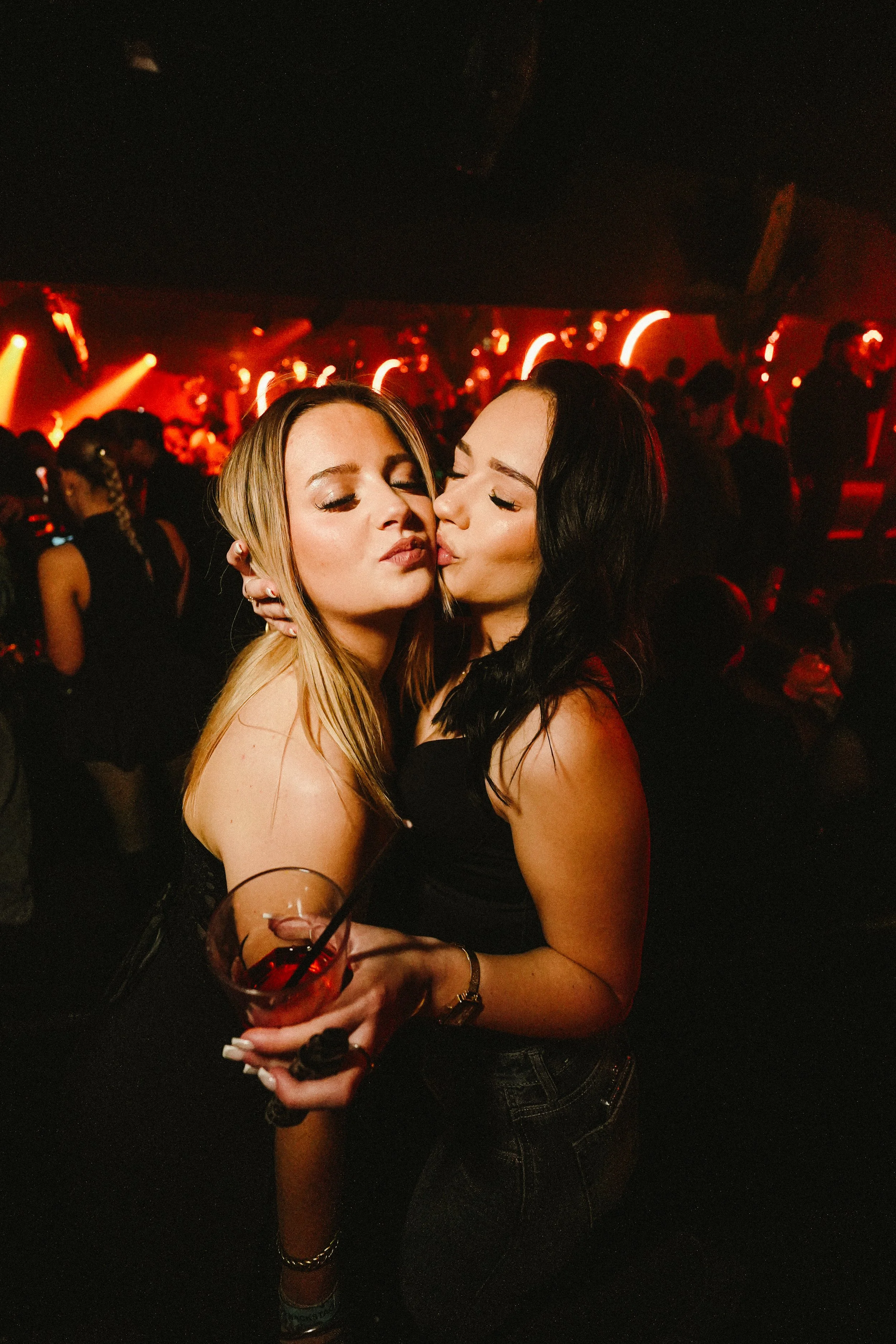 Two women embrace and kiss at a dimly lit nightclub or party, with other people dancing and red lighting in the background.