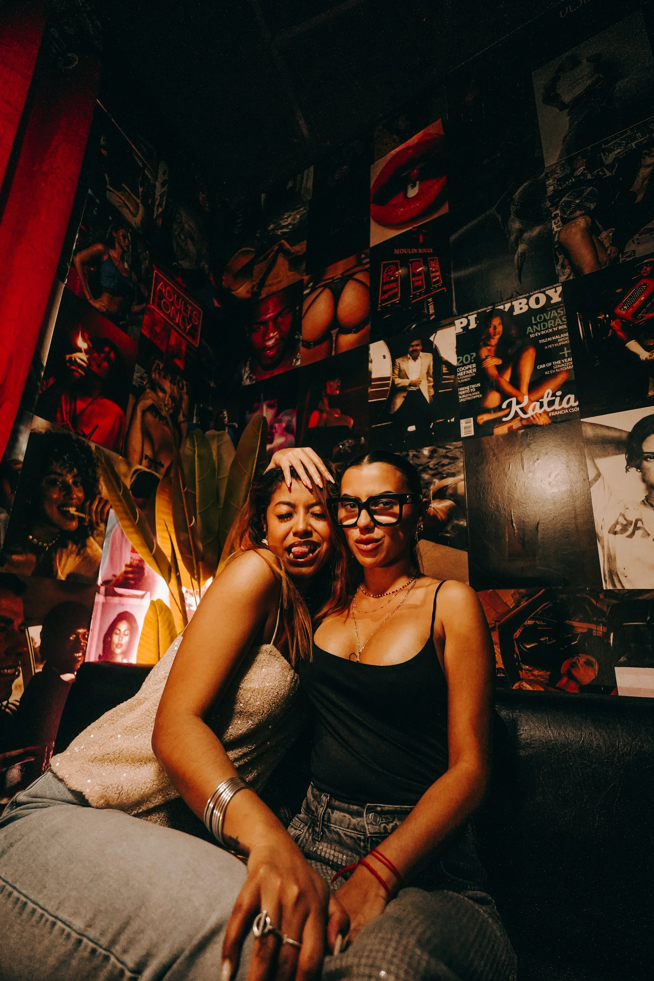 Two women sitting close together in front of a wall decorated with various posters and photographs, with their heads touching.