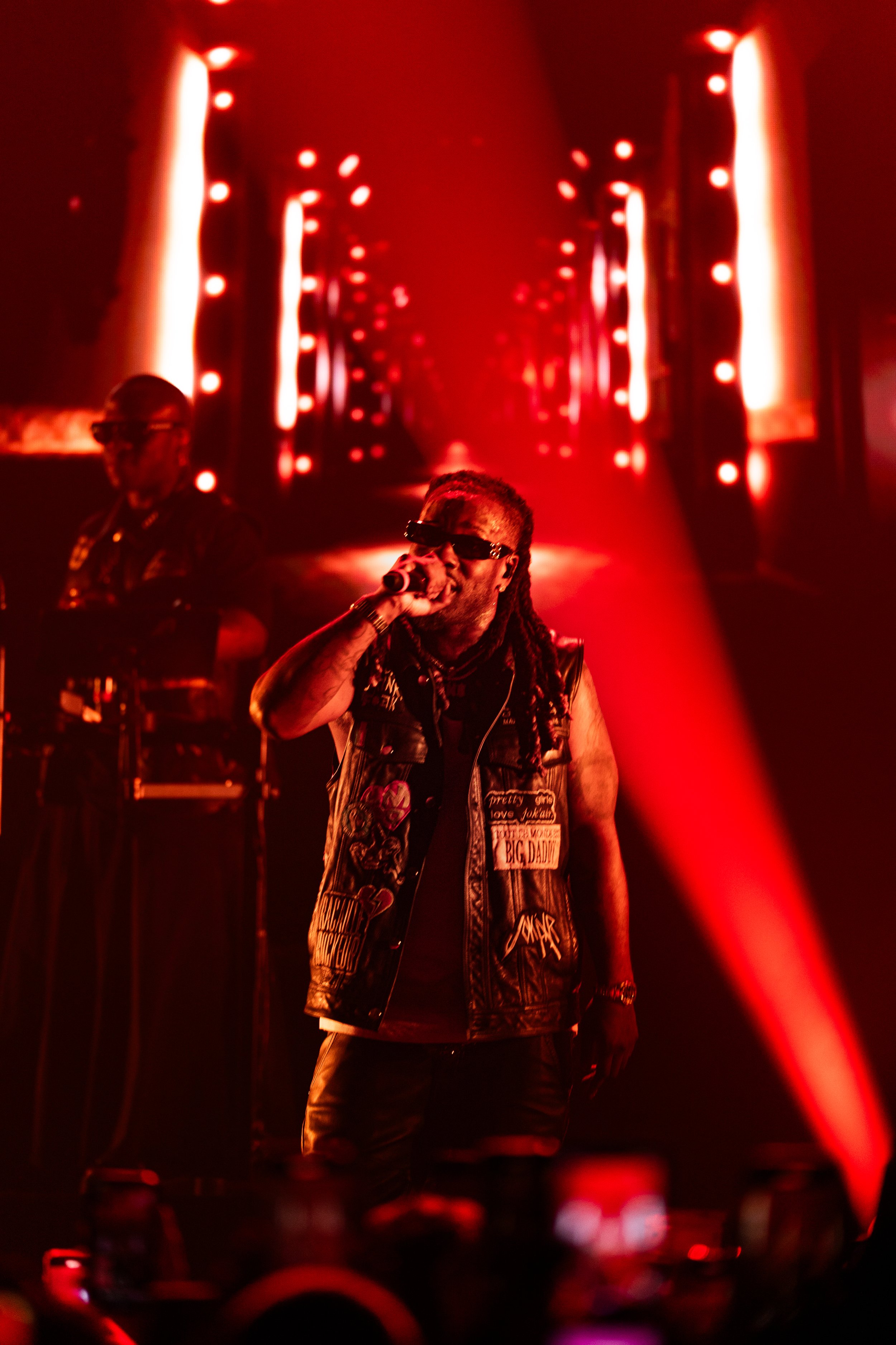 A male singer with long dreadlocks wears sunglasses and a black sleeveless vest with various patches and patches, performing on stage with a microphone. In the background, a DJ in a hat and headphones operates turntables. The stage is illuminated with red lights and LED panels creating a vibrant atmosphere.
