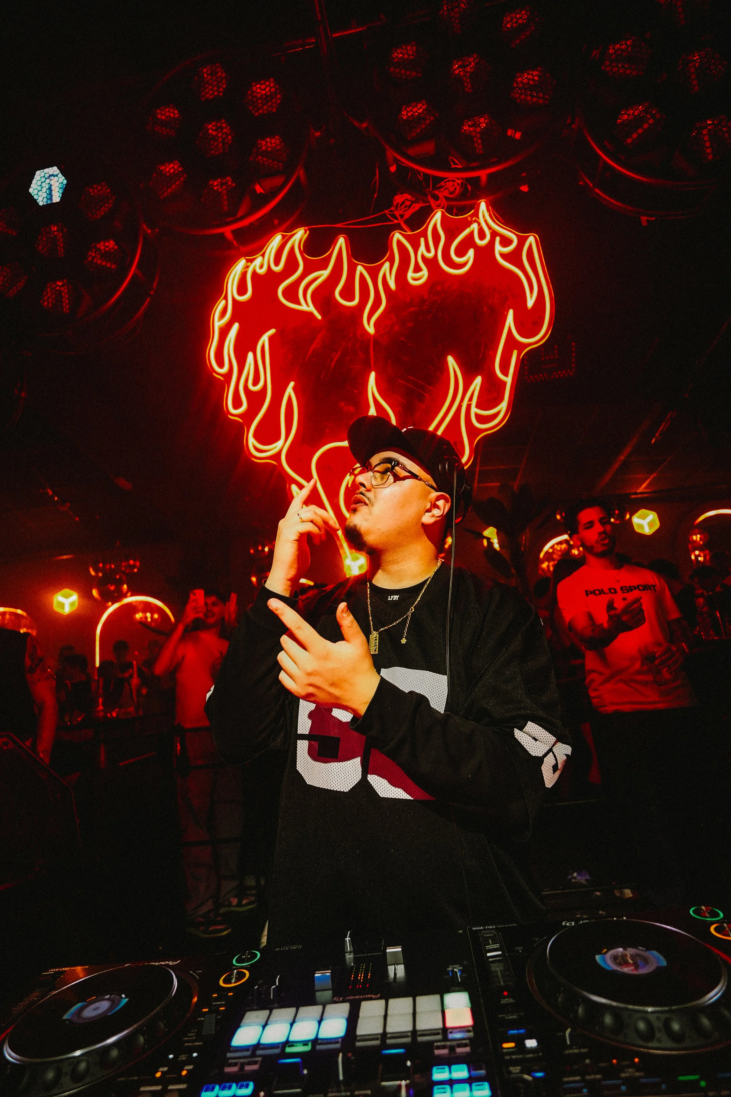 A DJ wearing glasses, a black cap, and a black jacket with white and red lettering, is standing at a DJ booth with colorful controls, in a nightclub with red lighting. Behind him, a red neon sign shaped like a flaming heart and a stylized face hangs on the wall, with several people in the background enjoying the music.