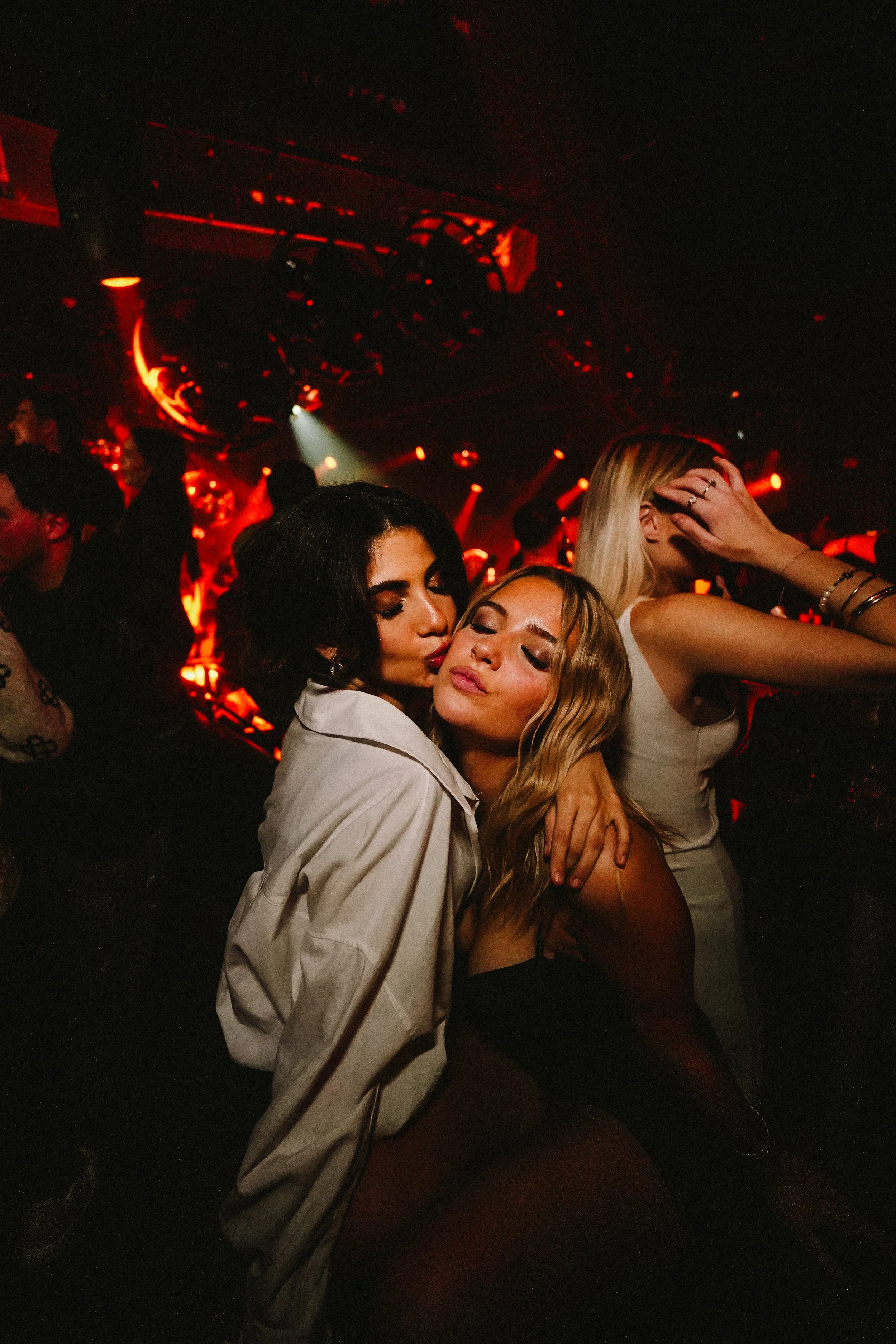 Two women are dancing closely together in a nightclub with red lighting, surrounded by other people, and a dark background with stage lights.