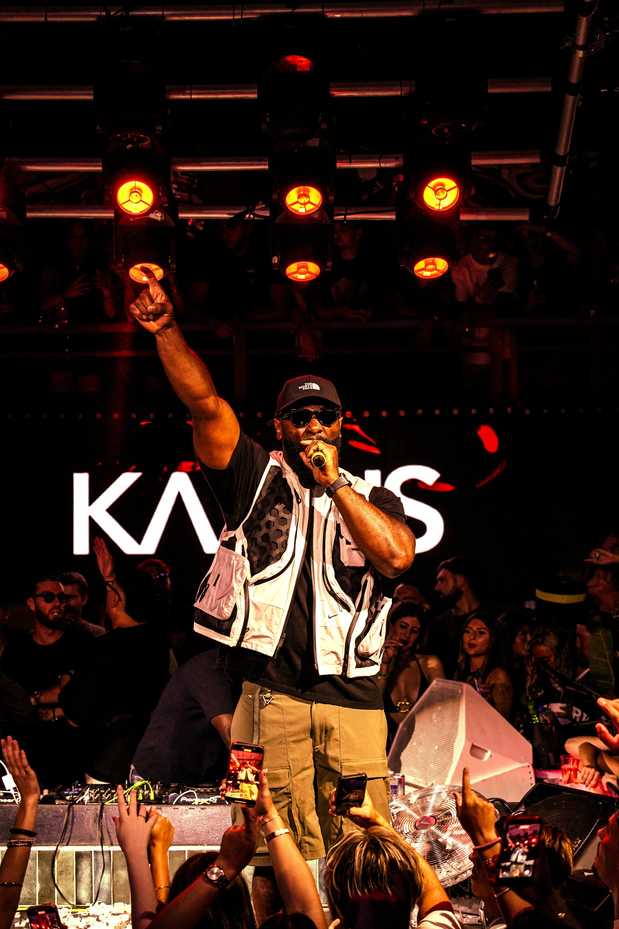 A male performer wearing sunglasses, a black cap, and a beige vest singing into a microphone on stage, with a crowd of people taking photos and cheering in the background, illuminated by red stage lights.