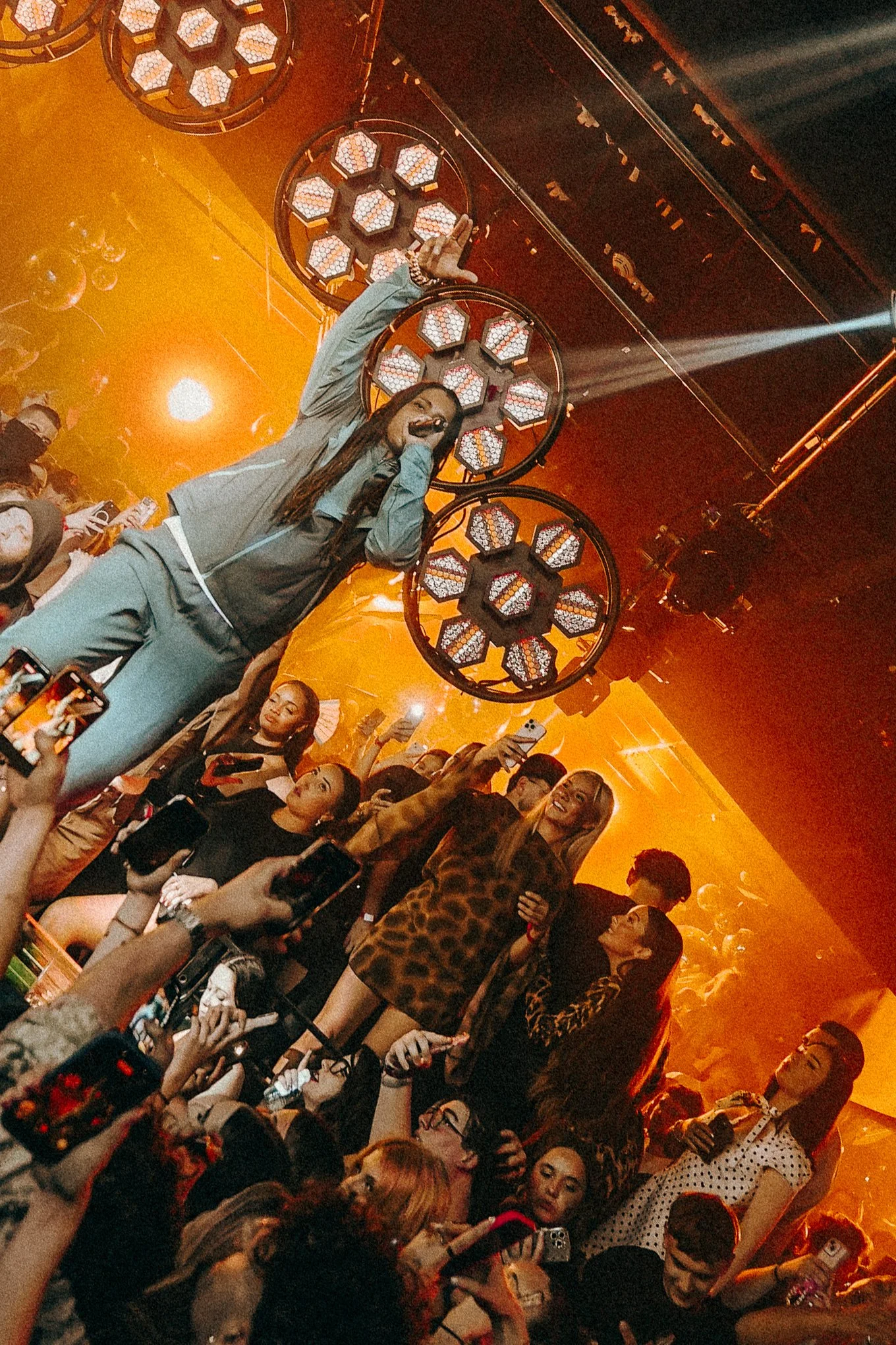 A woman on stage holding a microphone and gesturing to crowd. The stage has large, circular light fixtures overhead. Audience members are taking photos and videos, with some wearing patterned clothing and glasses. The lighting is warm with an orange hue.