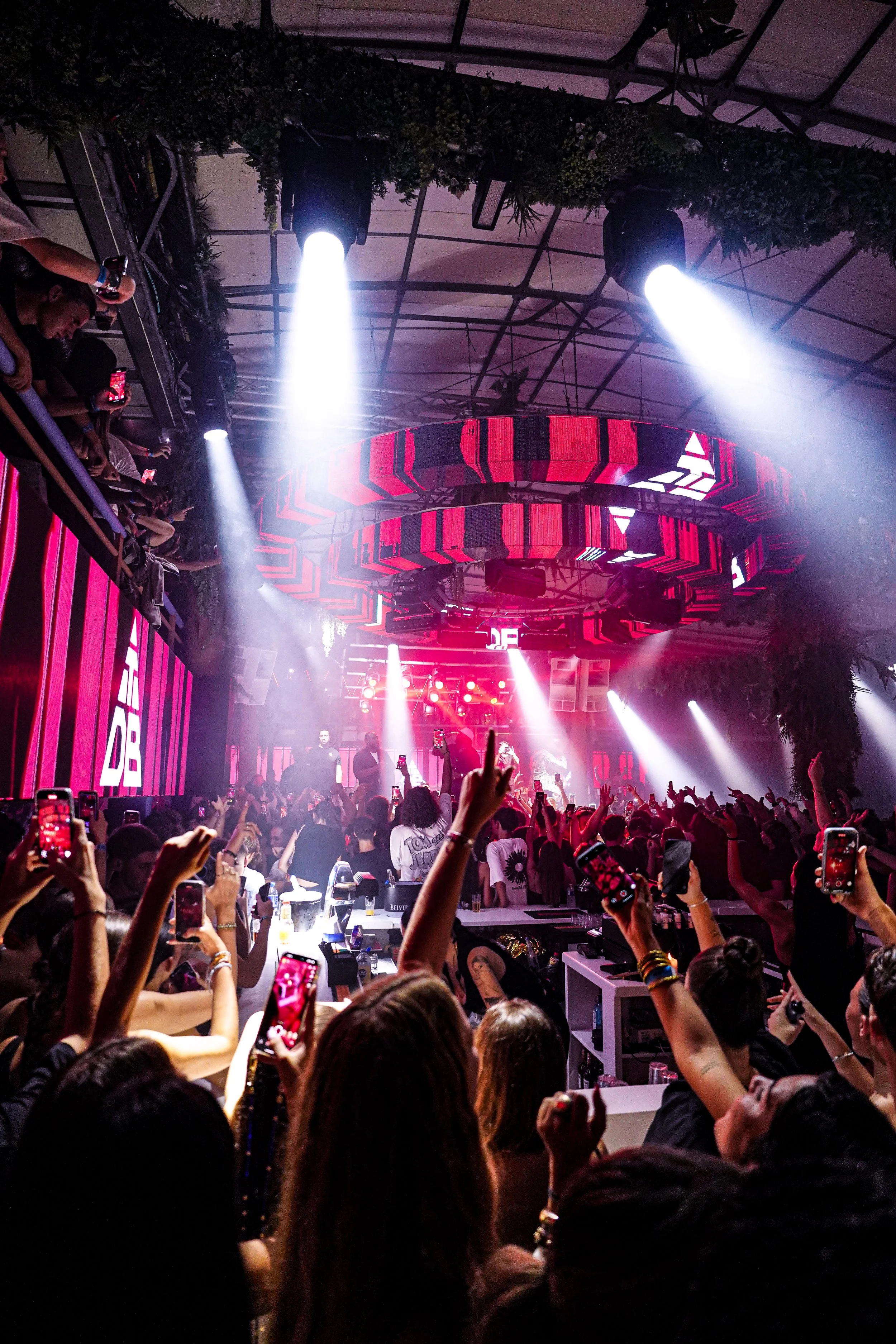 A crowded indoor concert or dance event with a stage, colorful lighting, and a large crowd of people, many of whom are taking photos or videos with their smartphones.