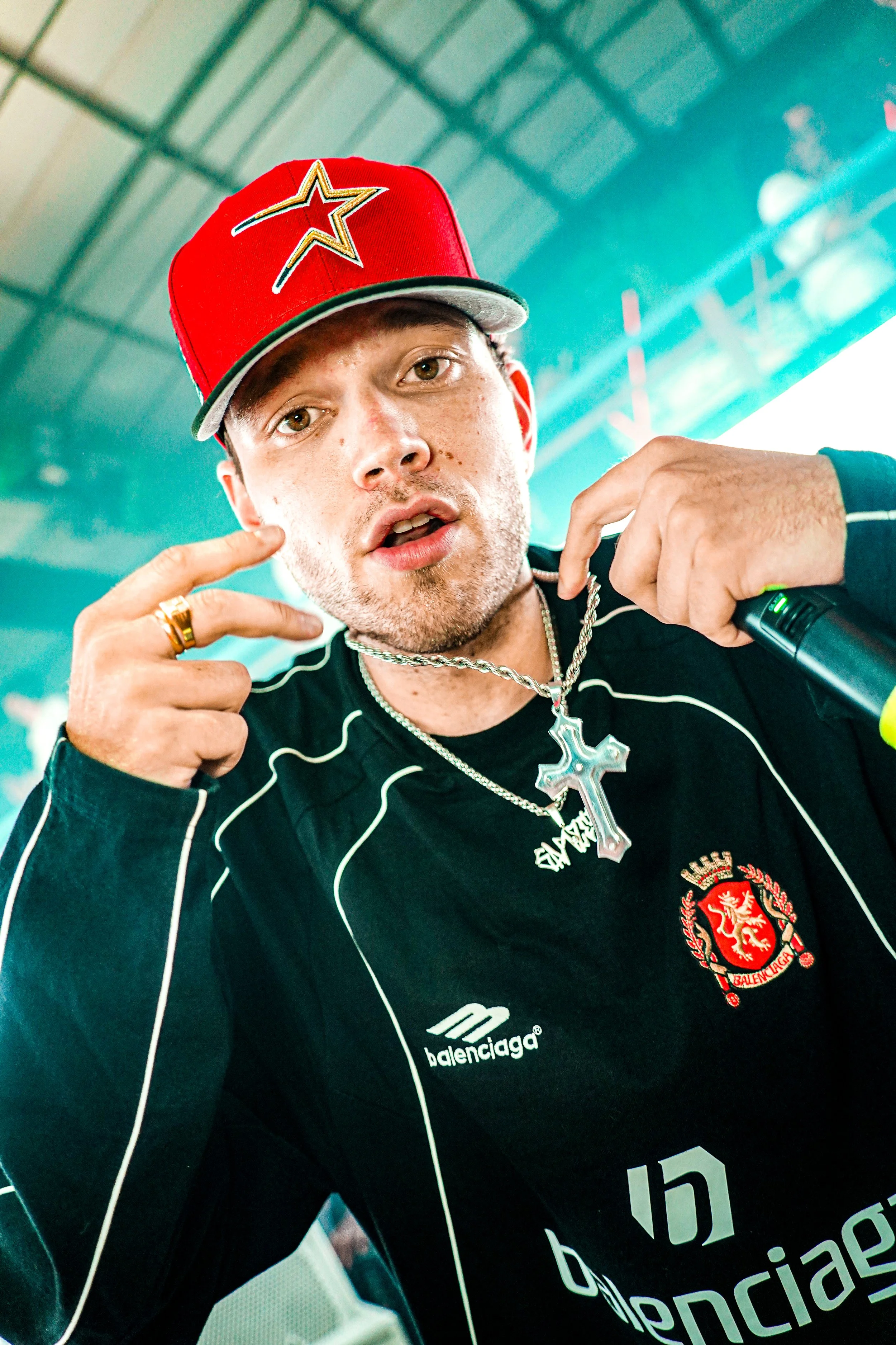 A young man wearing a red baseball cap with a star design, gold rings, a gold chain with a cross pendant, and a black Balenciaga shirt with white piping, is making a gesture near his cheek and looking at the camera.