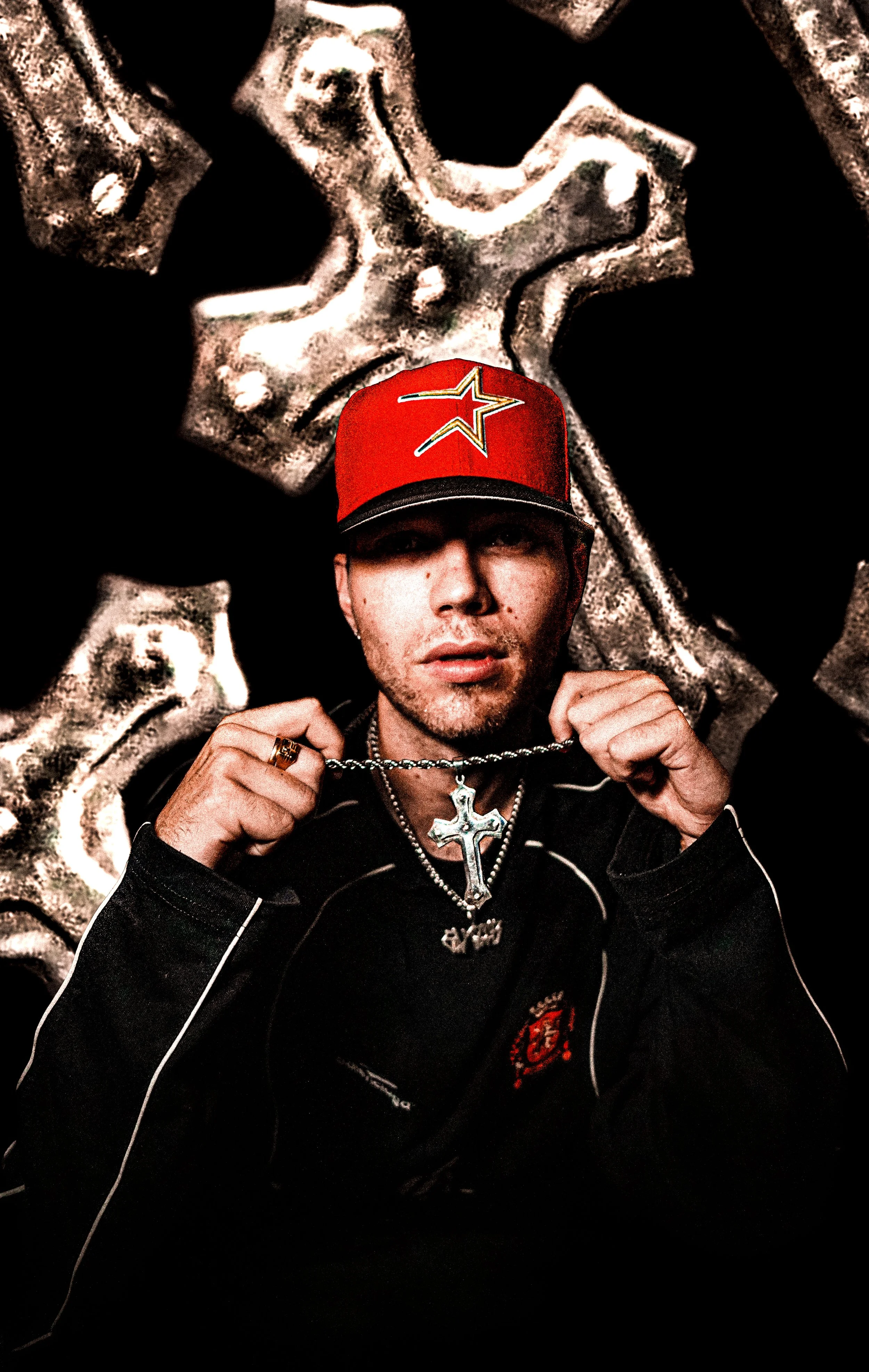 A man wearing a red baseball cap with a star logo, holding a chain with a cross pendant, posing in front of a textured metallic background.