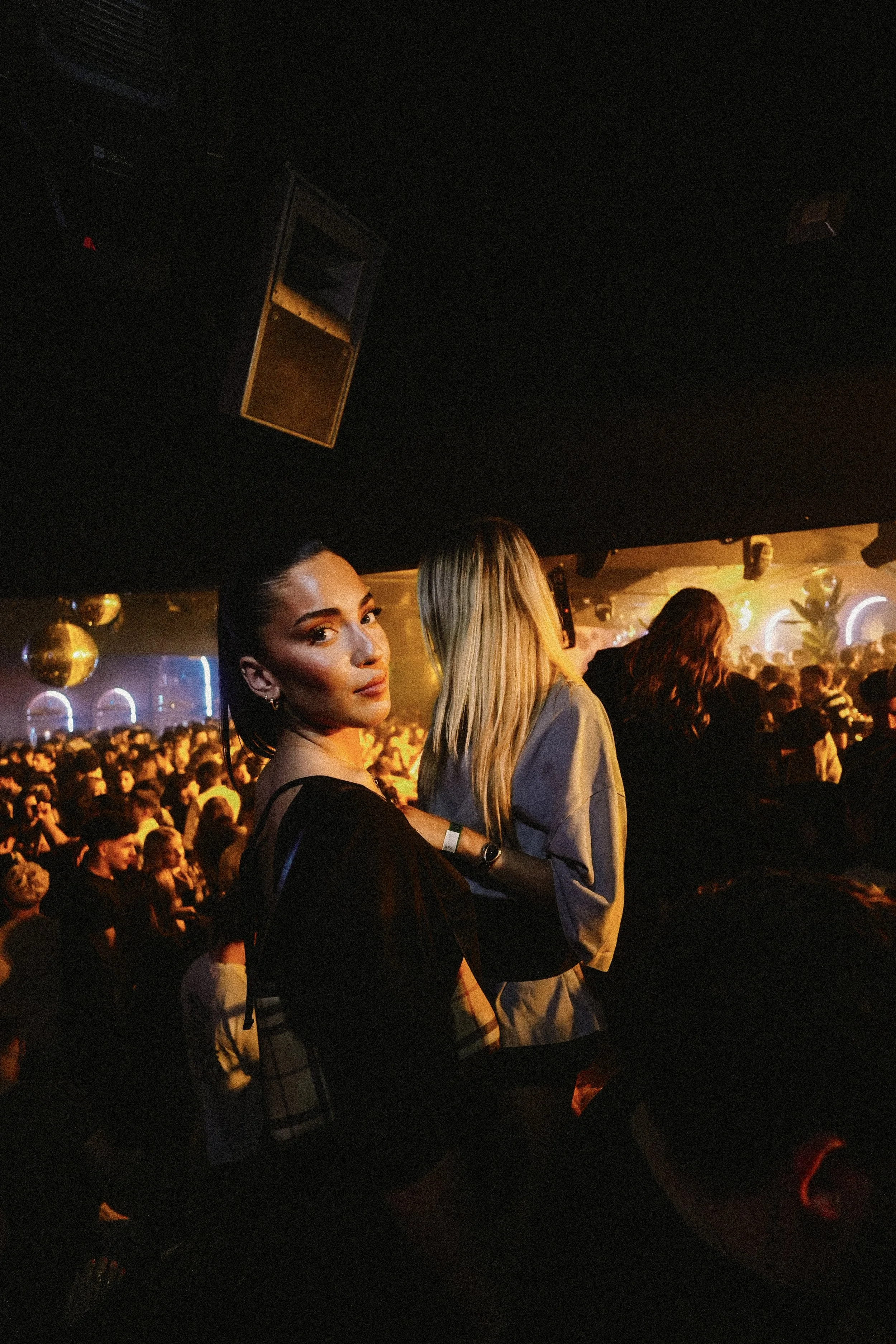 A young woman with dark hair and earrings looking at the camera in a crowded nightclub, with people dancing and colorful lights in the background.