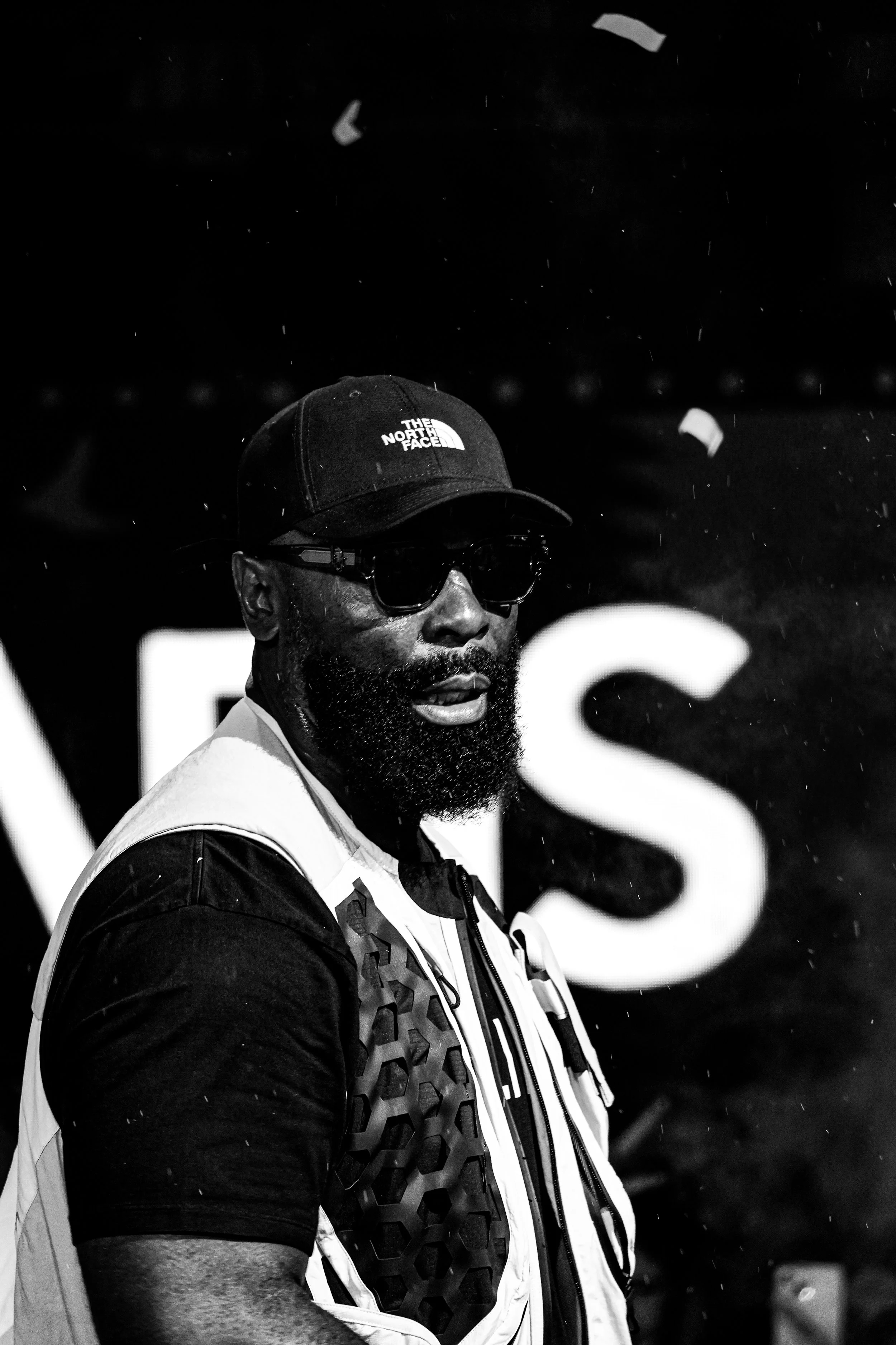 A man wearing a cap with 'The North Face' logo, sunglasses, and a vest, standing with a serious expression as snow falls around him during nighttime.