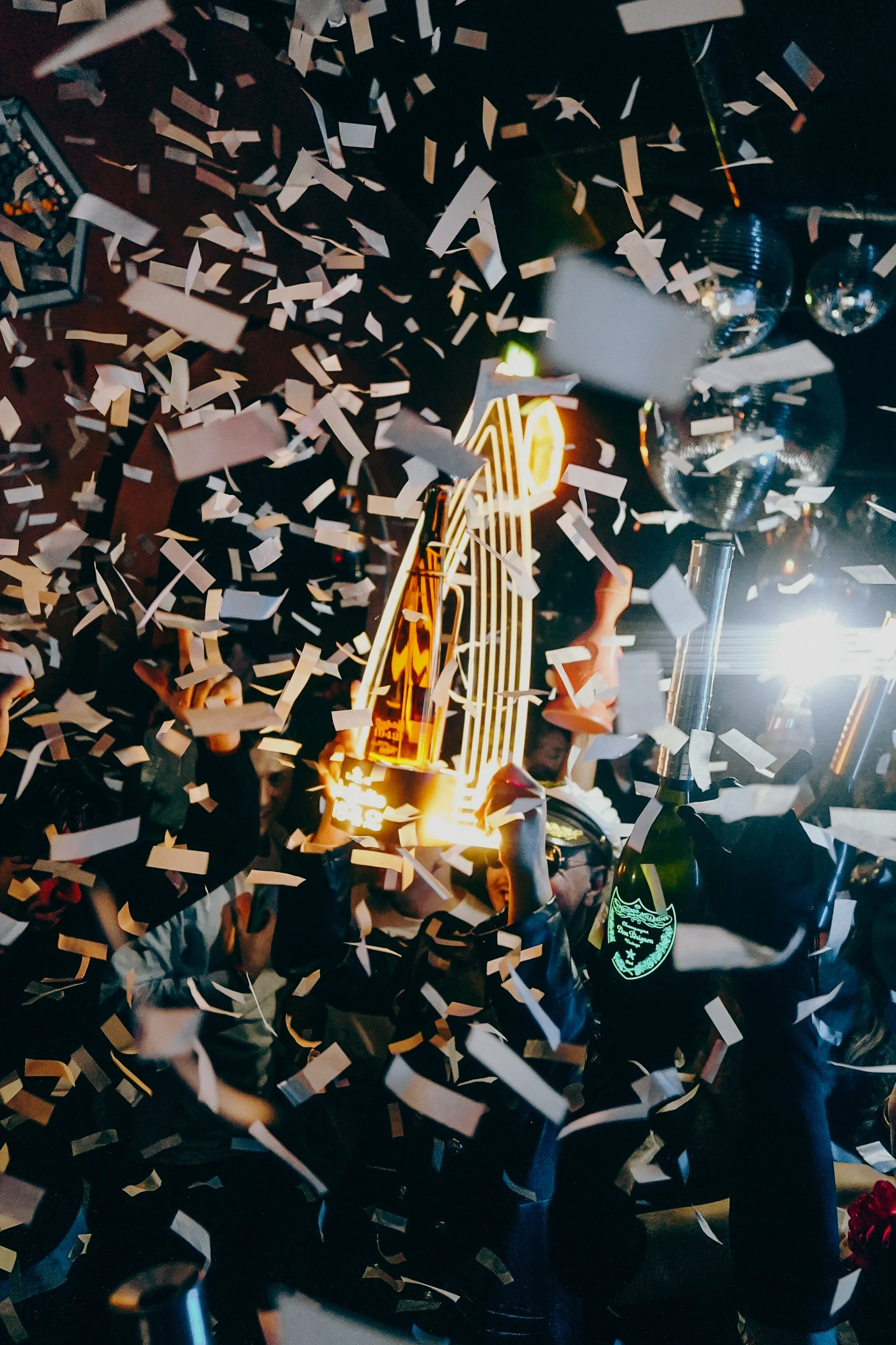 People celebrating a special occasion with confetti, holding a neon-lit sign, and glasses of champagne.
