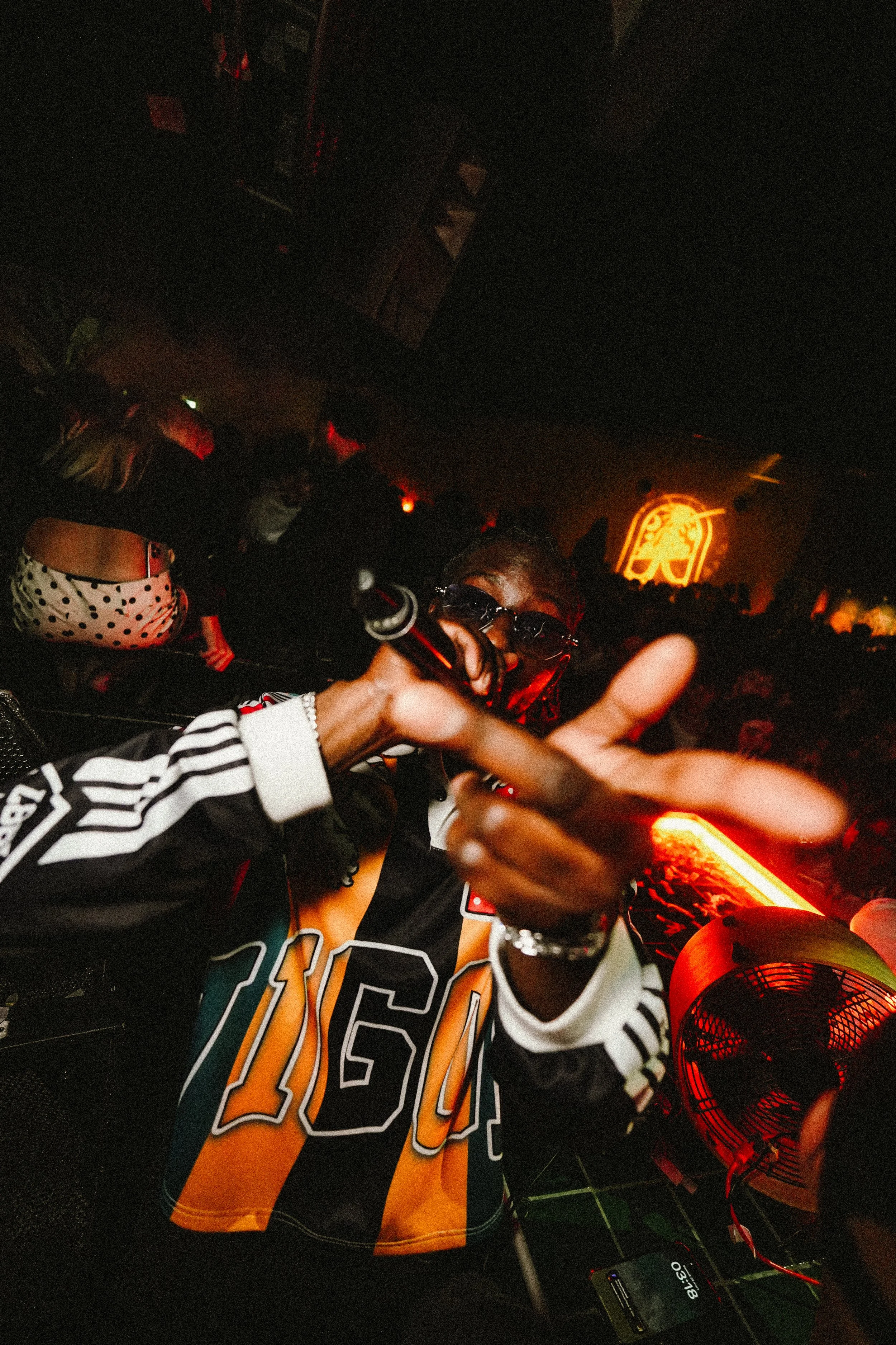 A person in a sports jersey with the number 46, wearing sunglasses, is holding a microphone and giving a thumbs-up in a dimly lit, crowded nightclub or party scene with a DJ setup and a neon logo in the background.